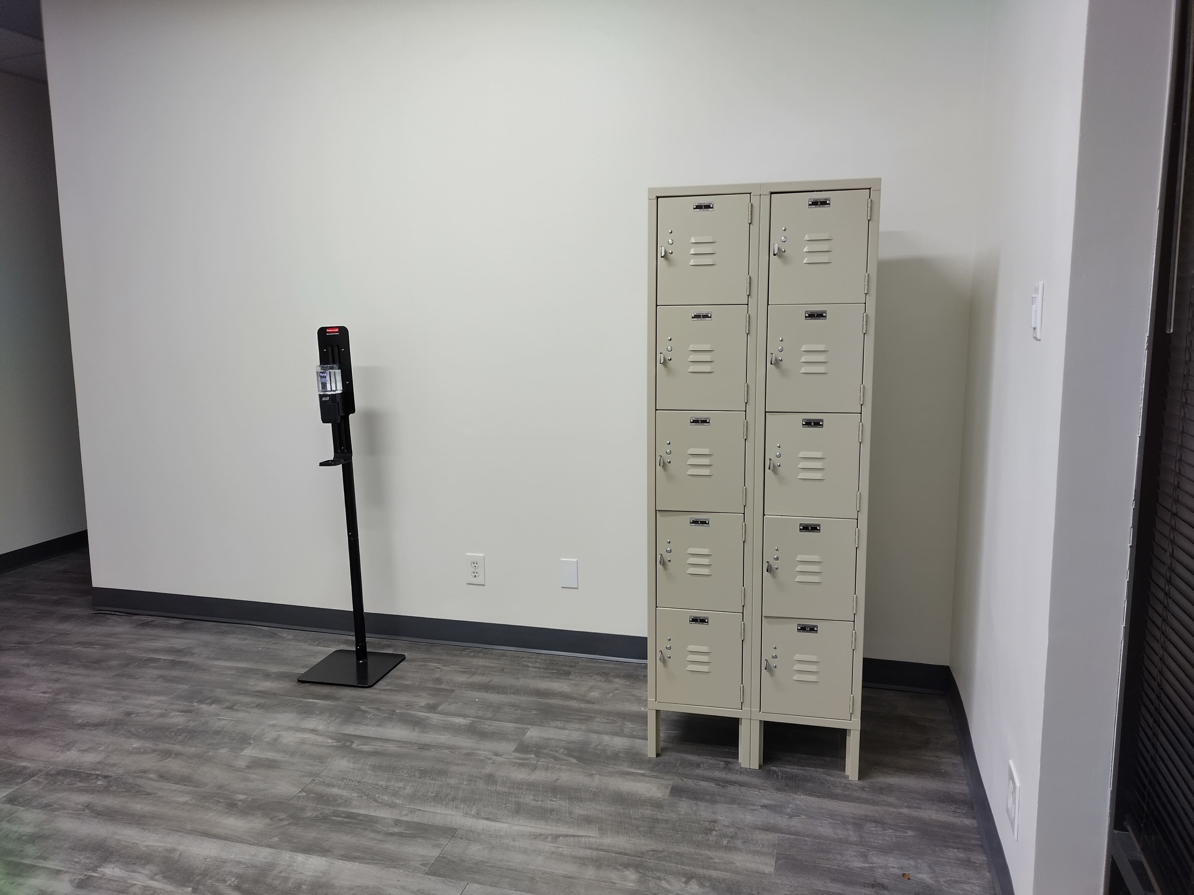 The image shows a small room with a metal locker unit on the right and a stand with what appears to be a hand sanitizer dispenser on the left.