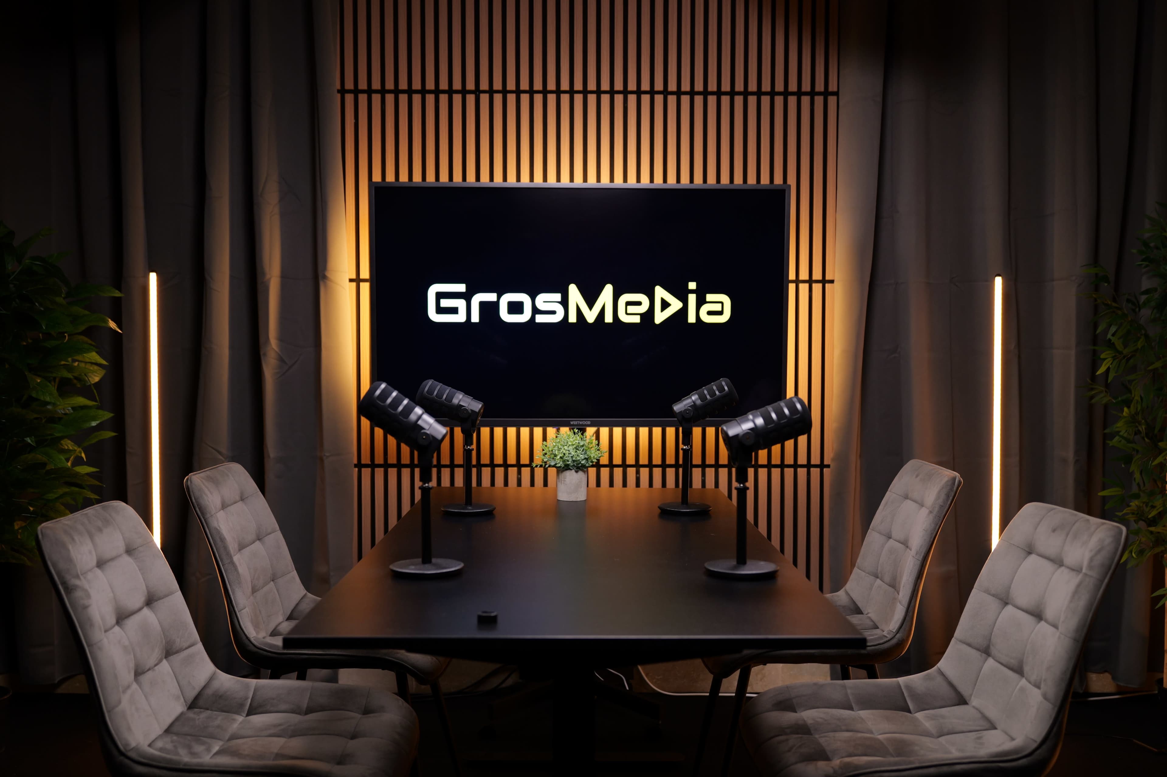 The image shows a modern conference room setup with a large screen displaying "GrosMedia," a black table, and several ergonomic chairs.