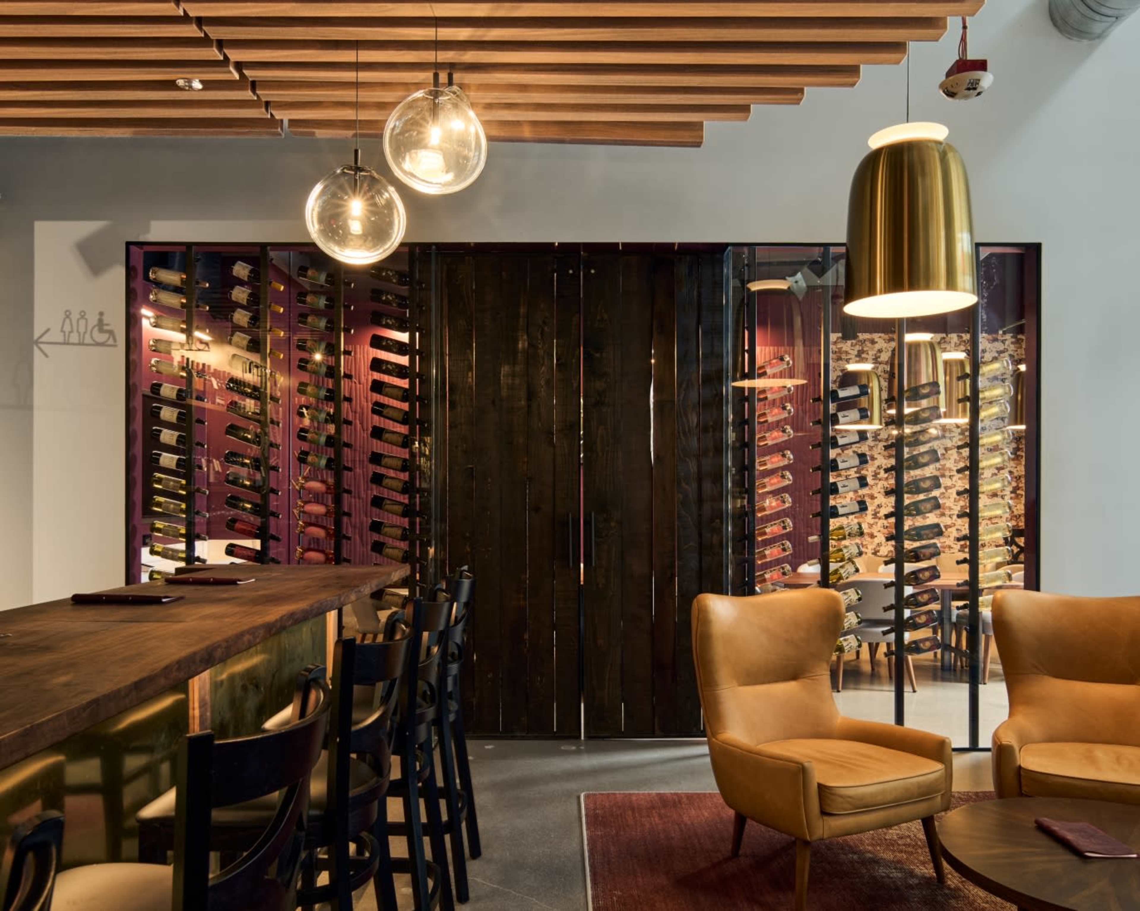The image shows a modern wine bar with a wooden counter, two stylish armchairs, and a wall display of wine bottles.