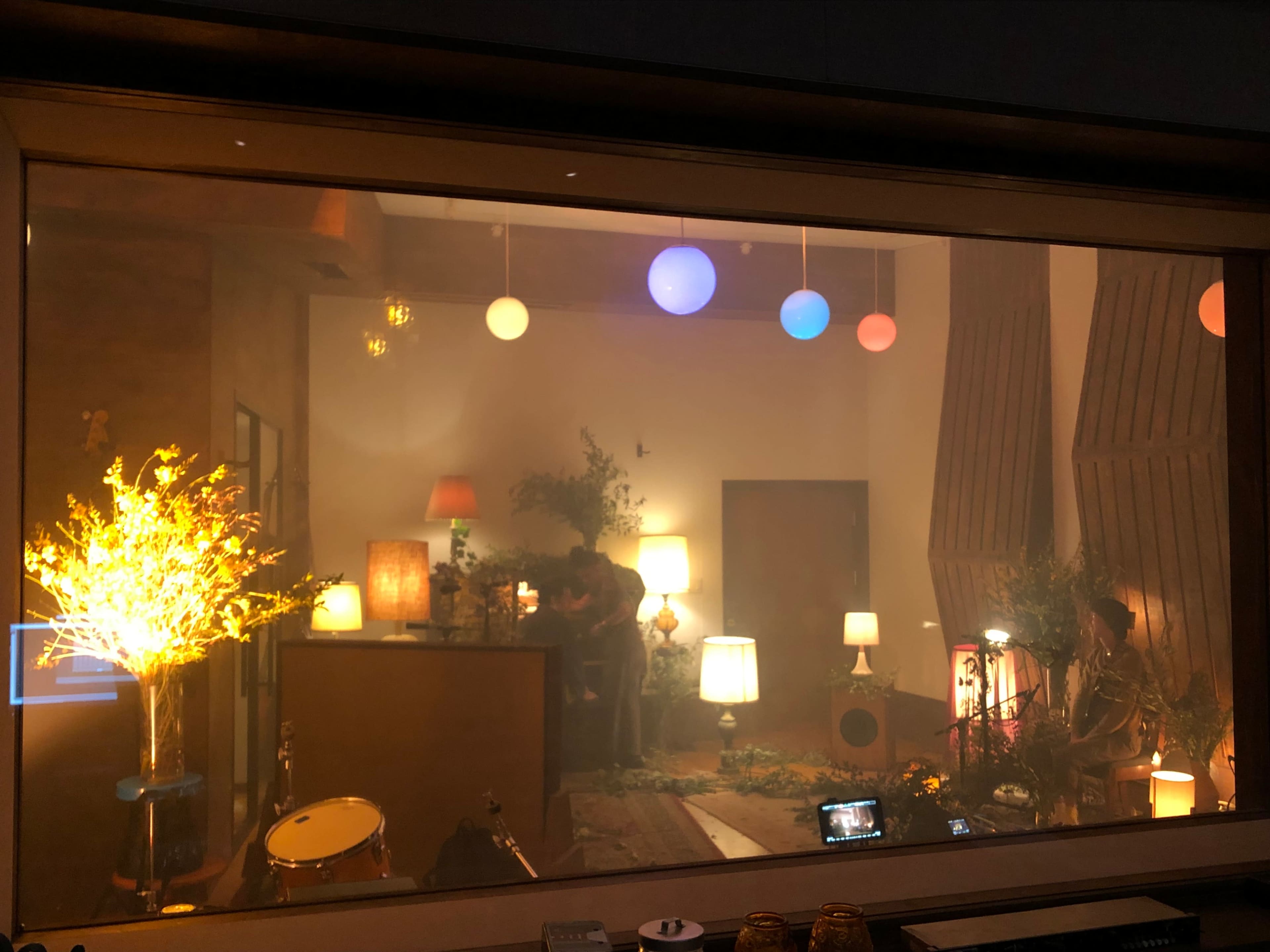 A studio room with various lamps, plants, and a set of drums, illuminated by colorful hanging lights.