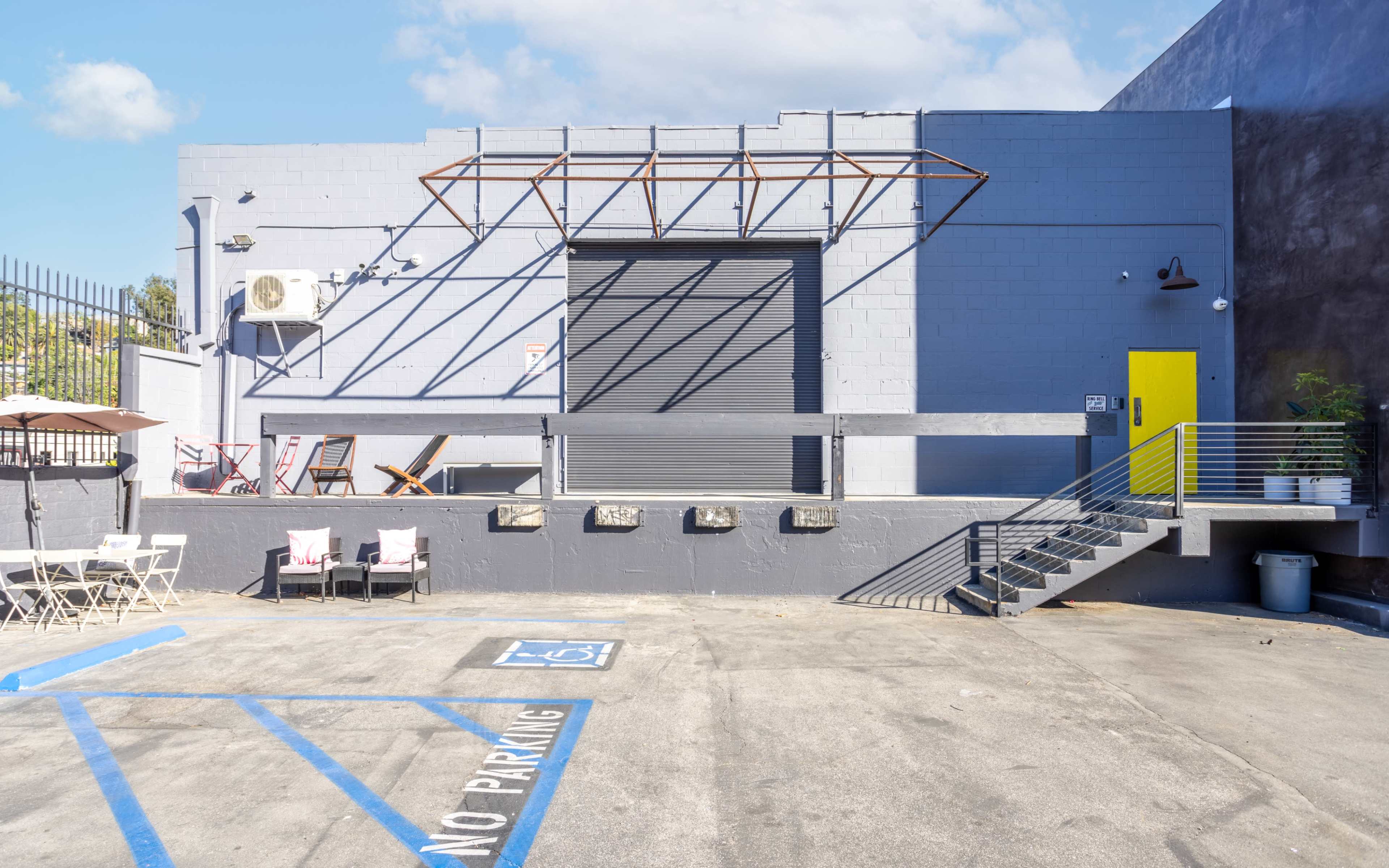 A gray building with a loading dock and a yellow door is set against a clear sky, featuring a parking area with marked spaces and no-parking signs.