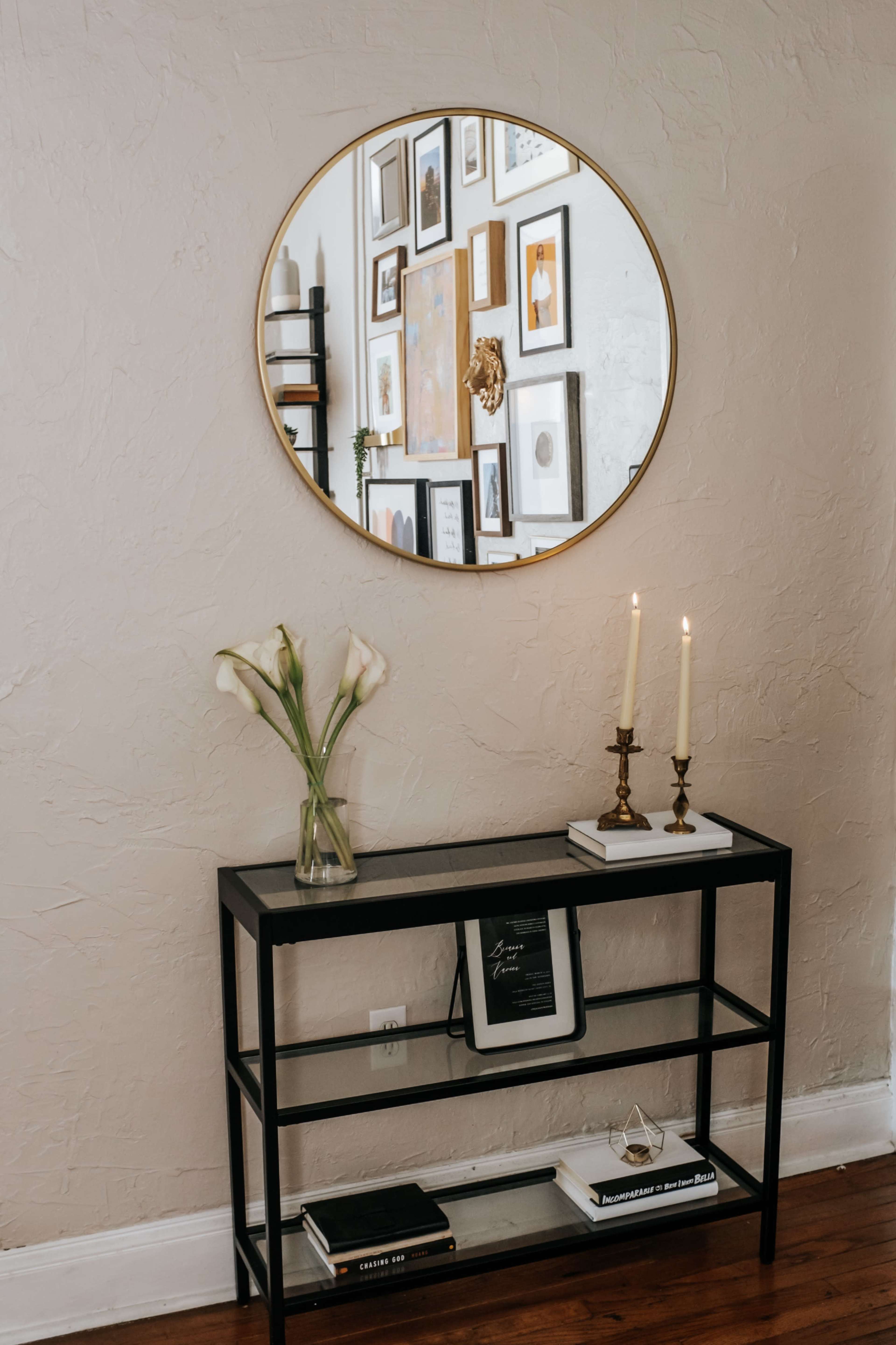 A modern black console table with a vase of flowers, candles, and books is positioned beneath a round mirror reflecting framed artwork on the wall.