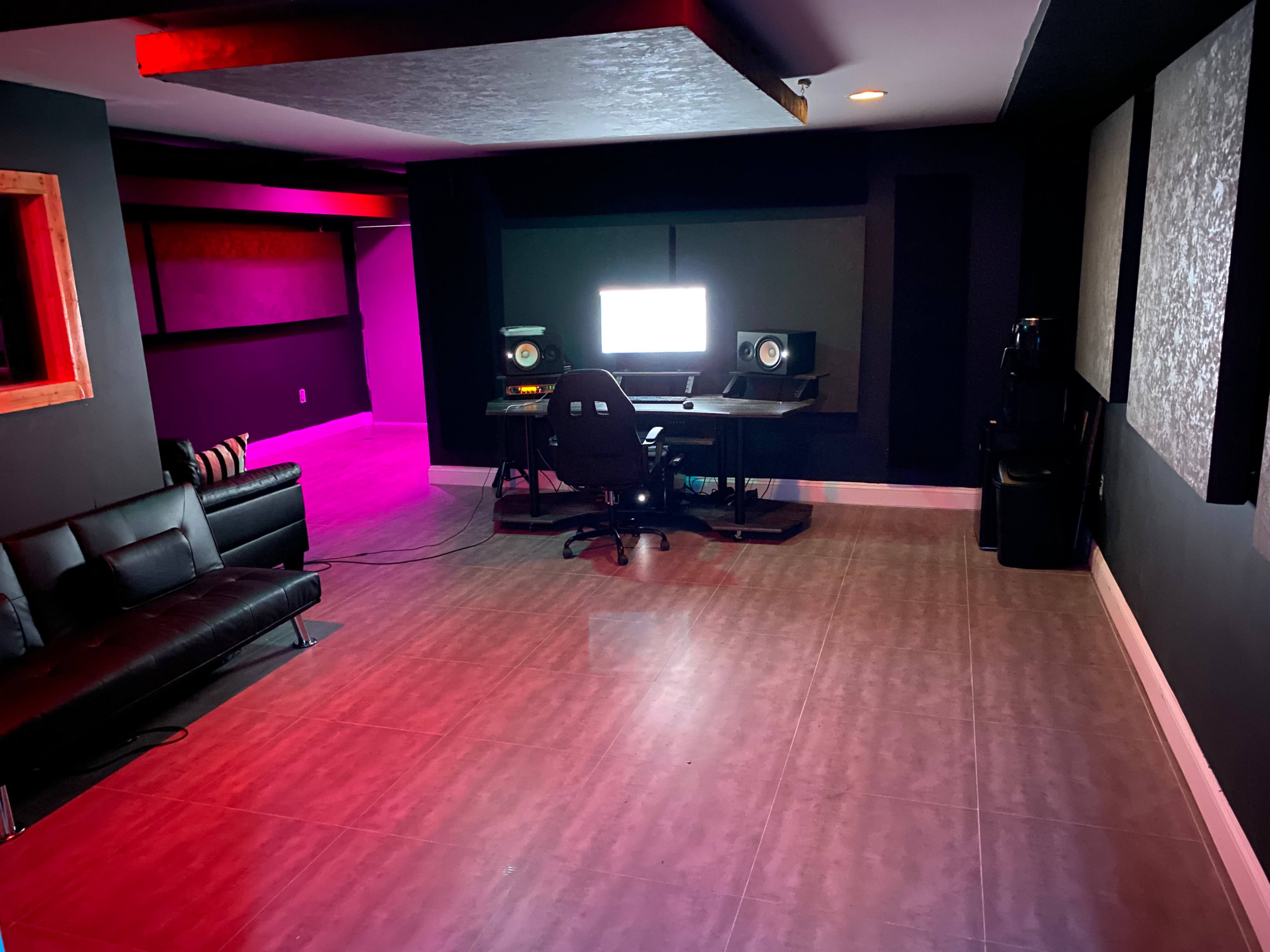 A modern music studio with a computer workstation, speakers, and a black sofa, illuminated by purple and red lighting.