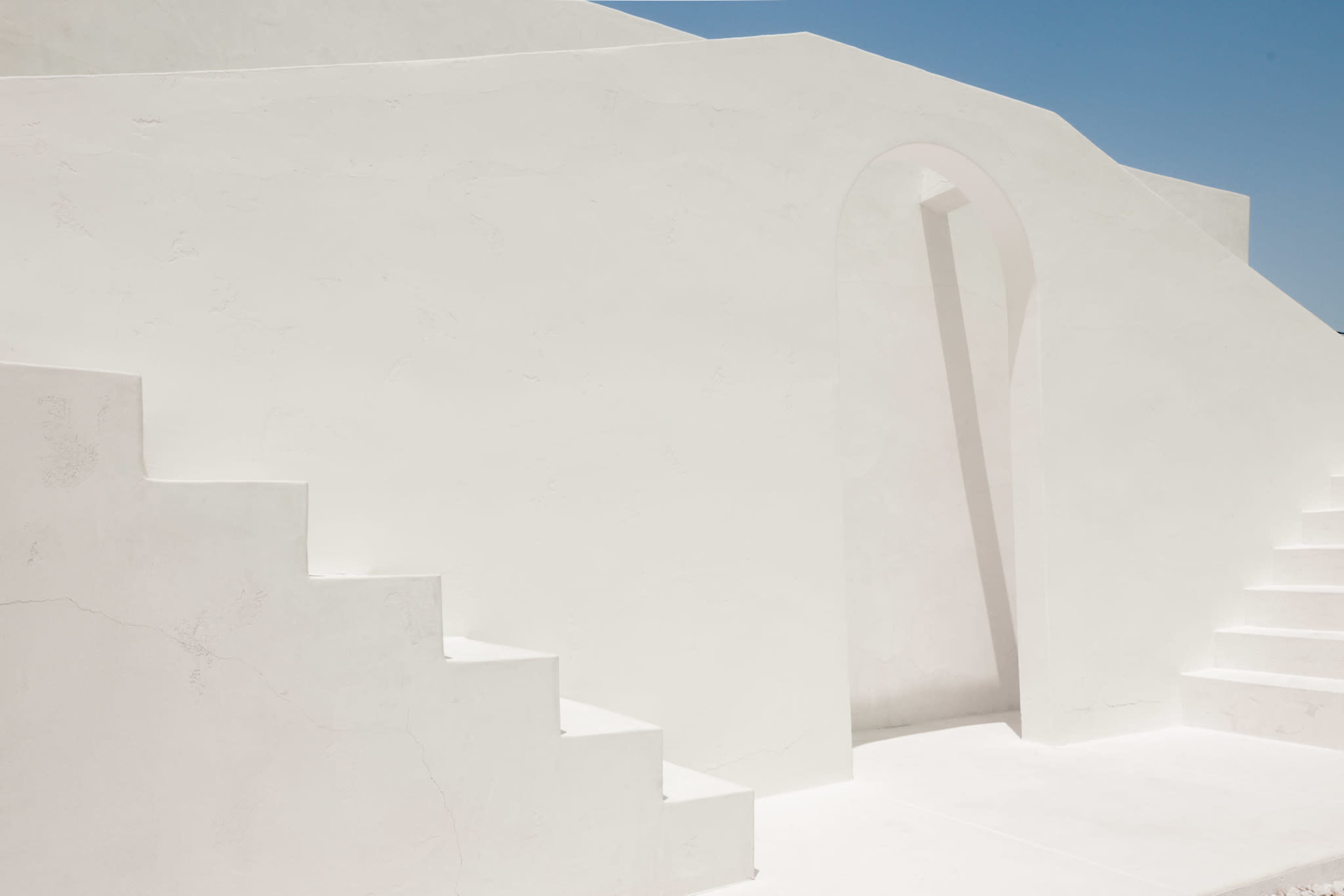 A minimalist architectural scene with white steps leading up to an archway framed by smooth, curved walls under a clear blue sky.