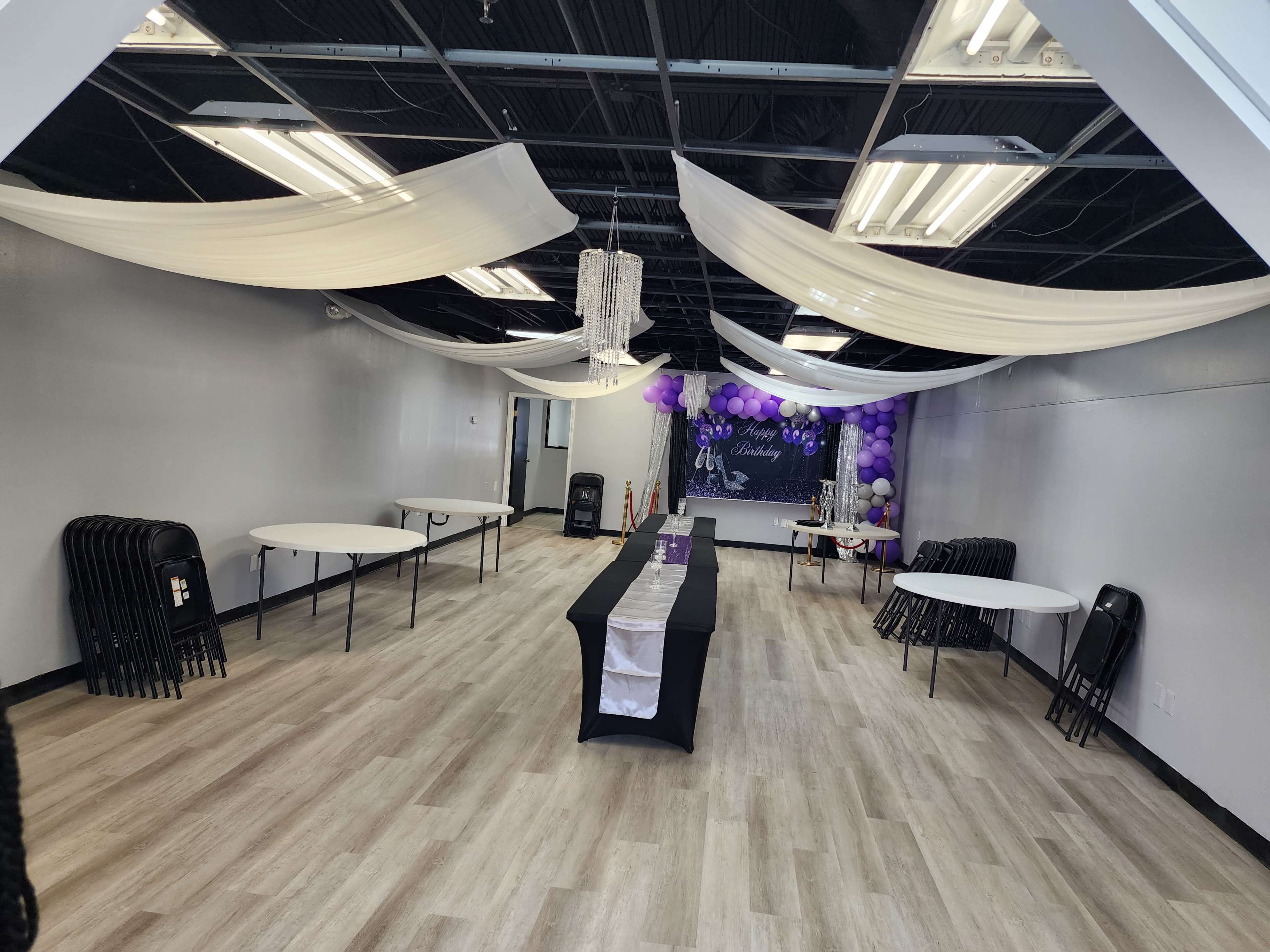 A spacious event room with several folding tables, draped fabric overhead, and decorations featuring purple balloons.