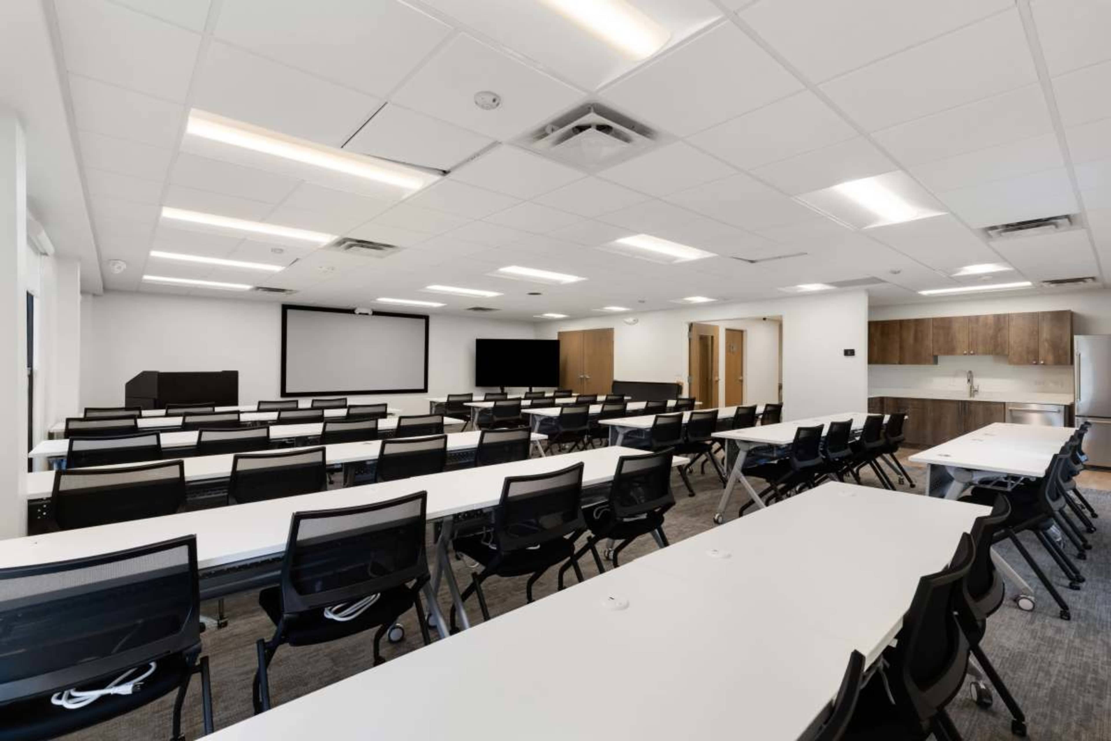 The image shows a modern conference room arranged with multiple rows of tables and chairs, featuring two large screens at the front and a kitchenette area in the back.