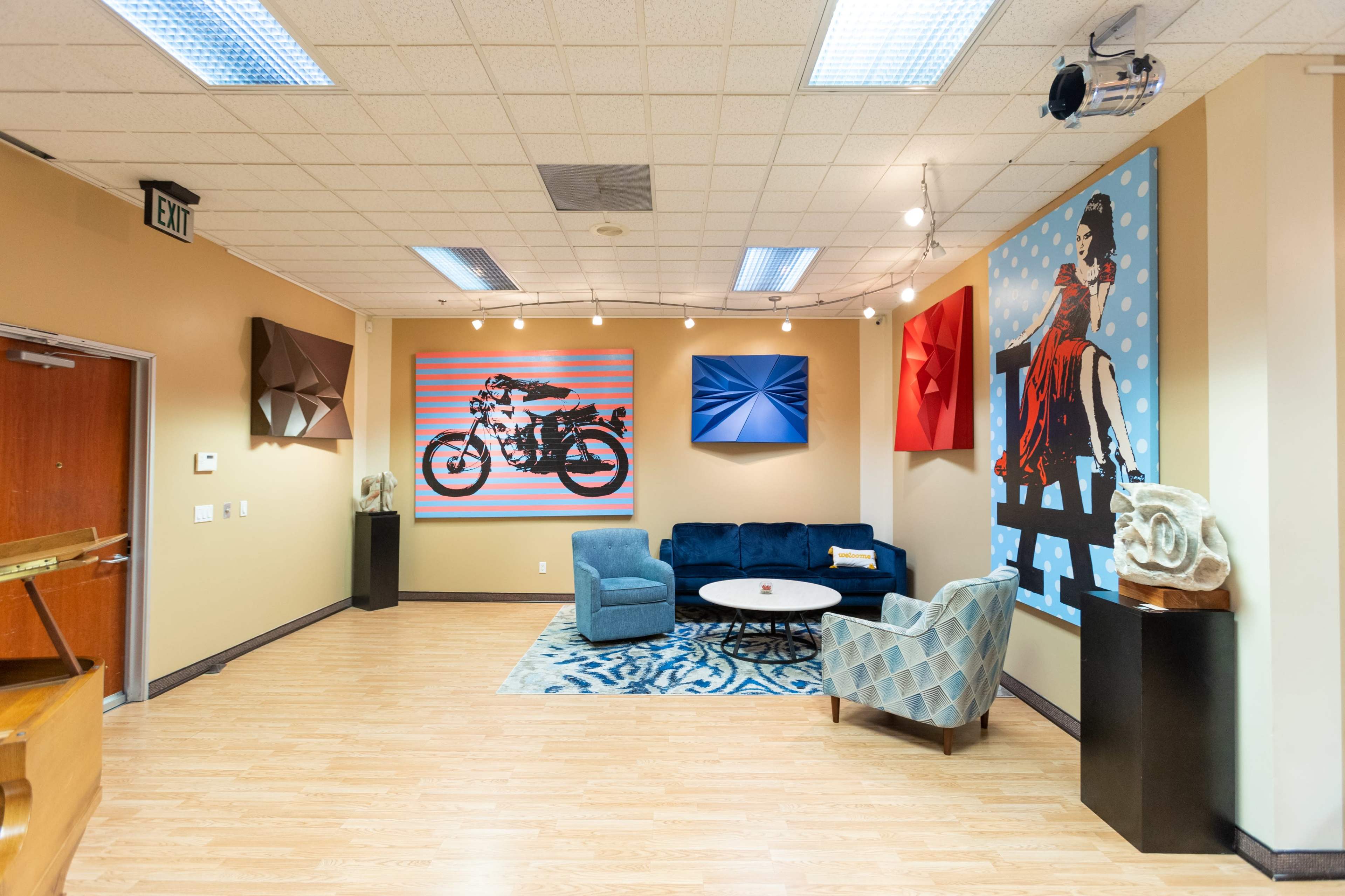A modern art gallery featuring a blue couch, a circular coffee table, and various colorful artworks on the walls, including a motorcycle and a figure in red.