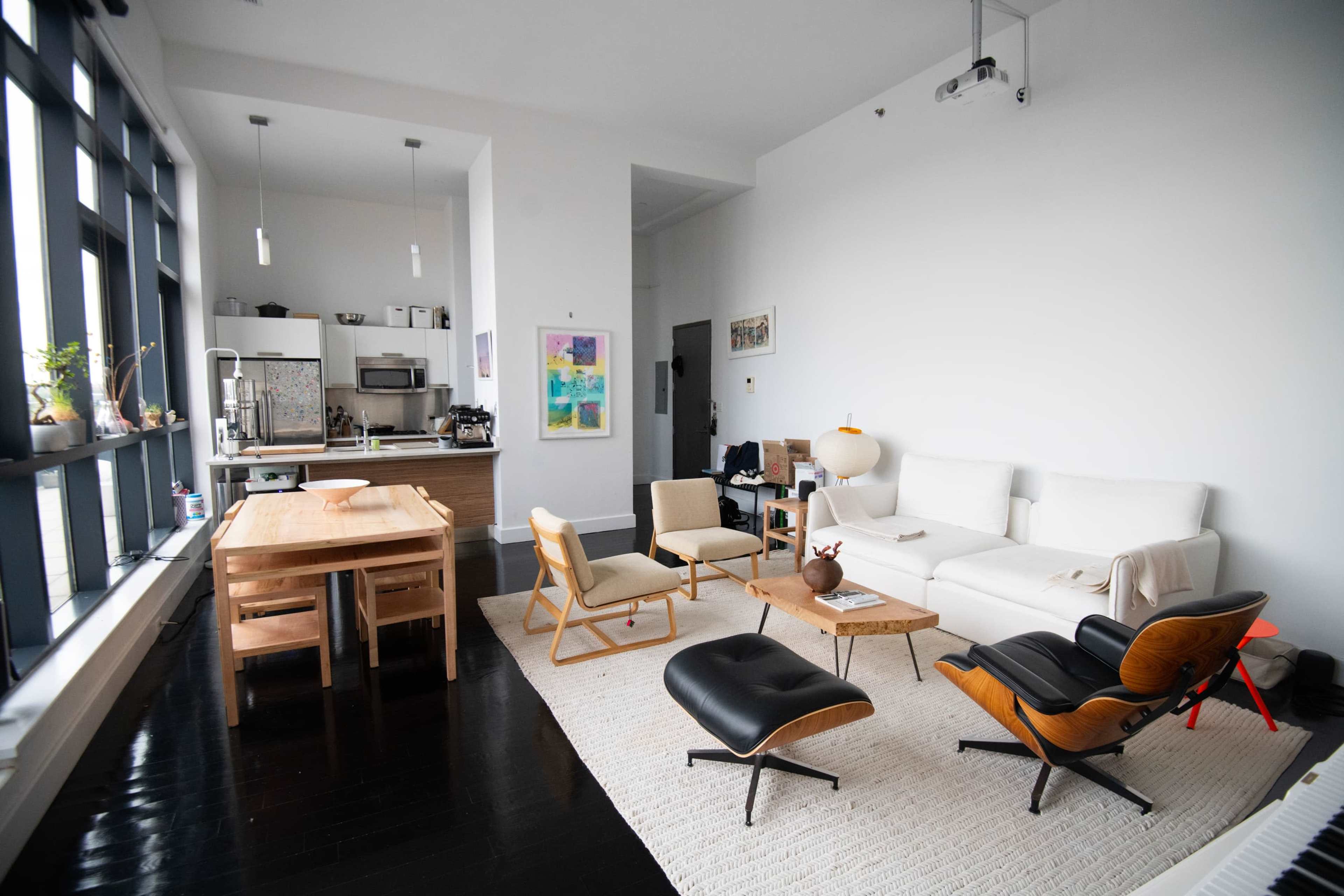 A modern living space features a white sofa, wooden dining table, and various seating arrangements with large windows allowing natural light.