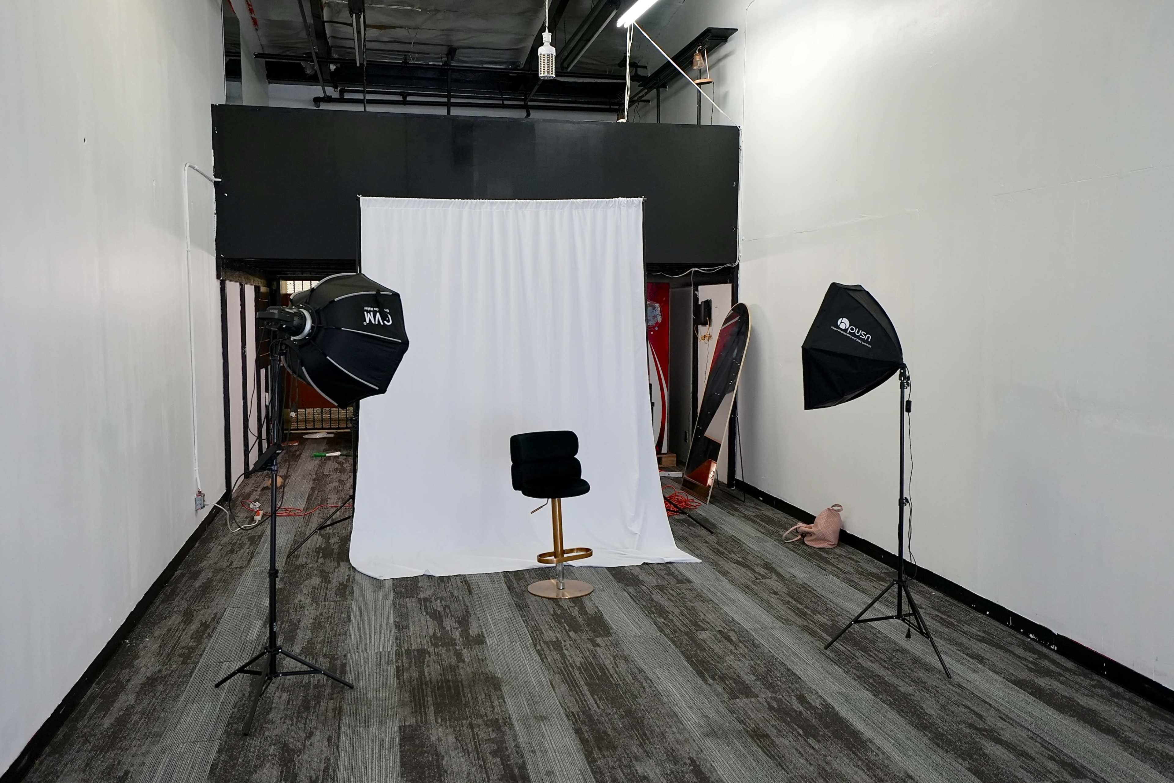 The image shows a photography setup consisting of a white backdrop, a stool, and two light sources positioned on stands in an empty, industrial-looking room.