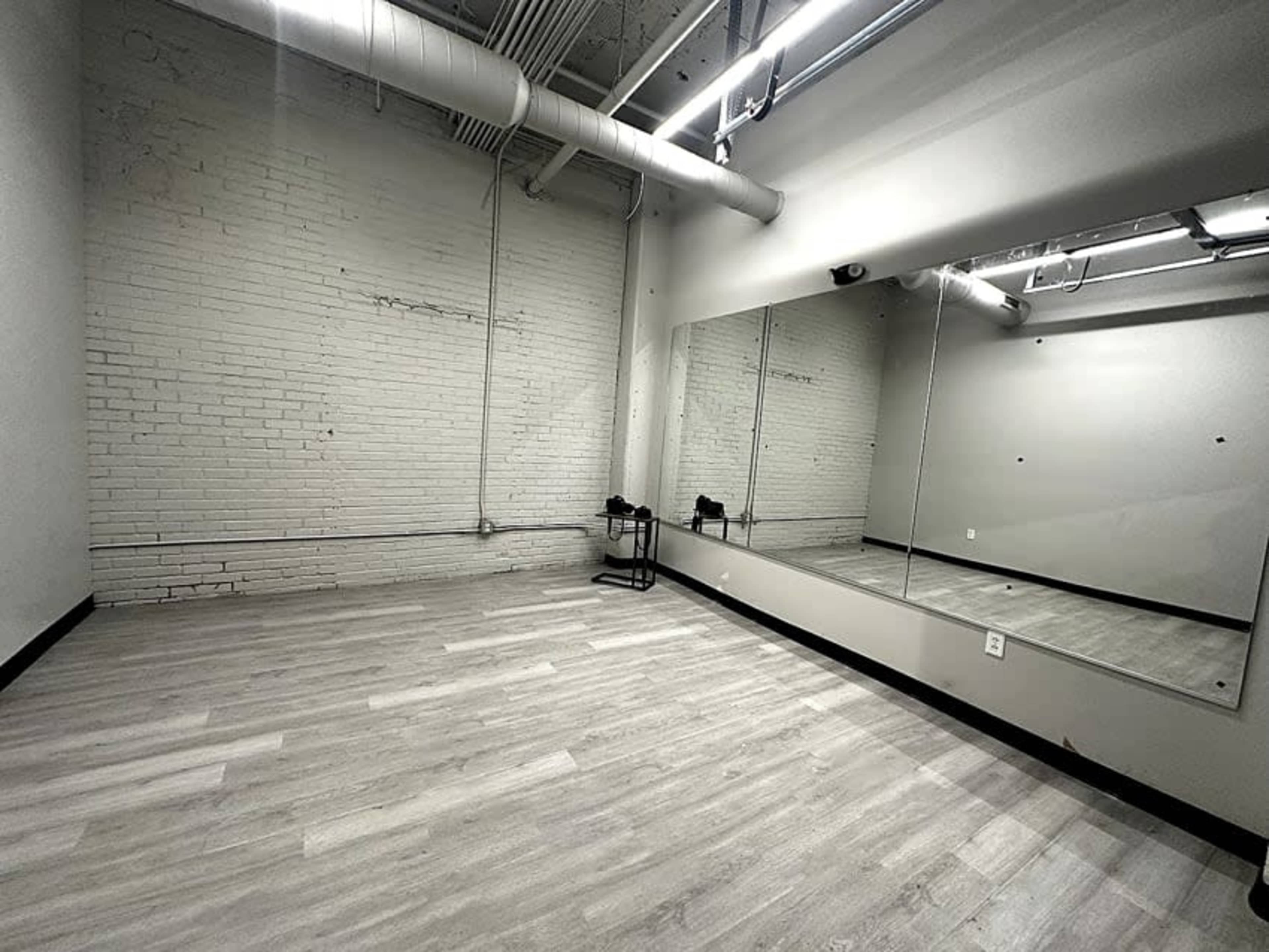 The image shows a spacious, empty room with gray wooden flooring, white brick walls, and a large mirror on one side.