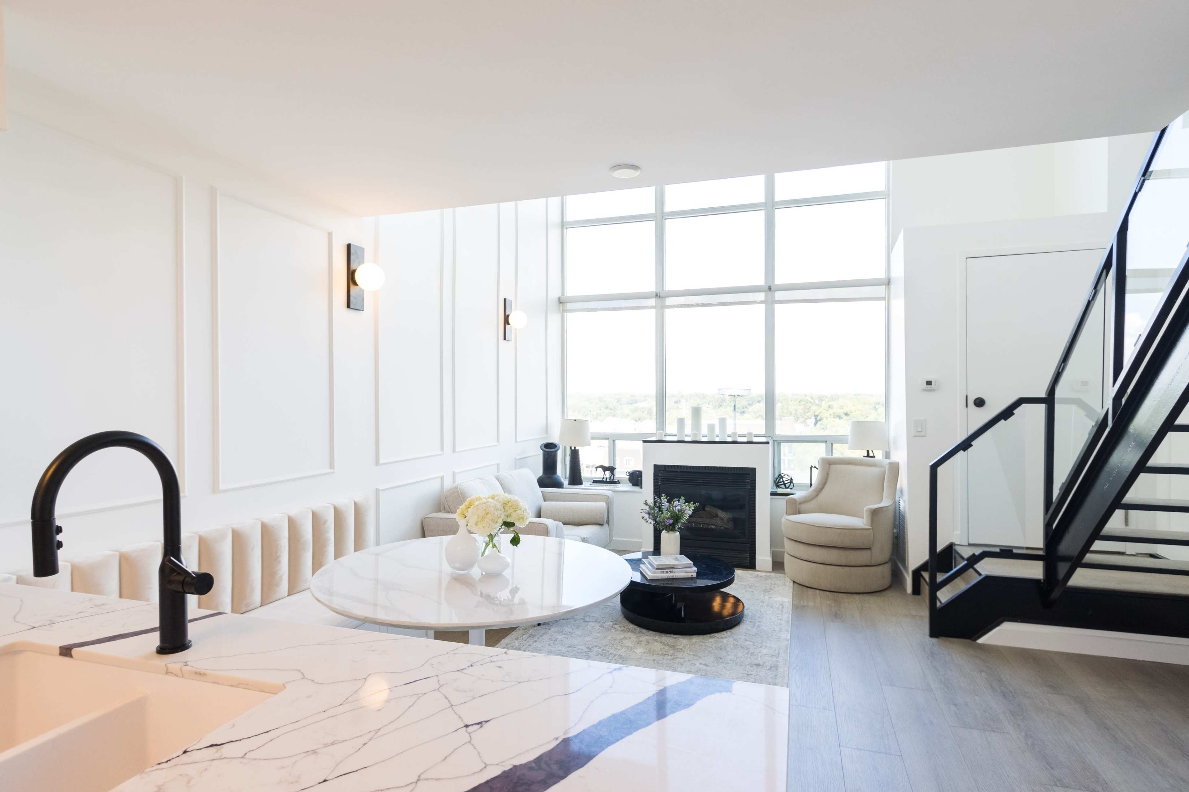 A modern living space features a marble dining table, white seating, a large window with a view, and a staircase leading to an upper level.