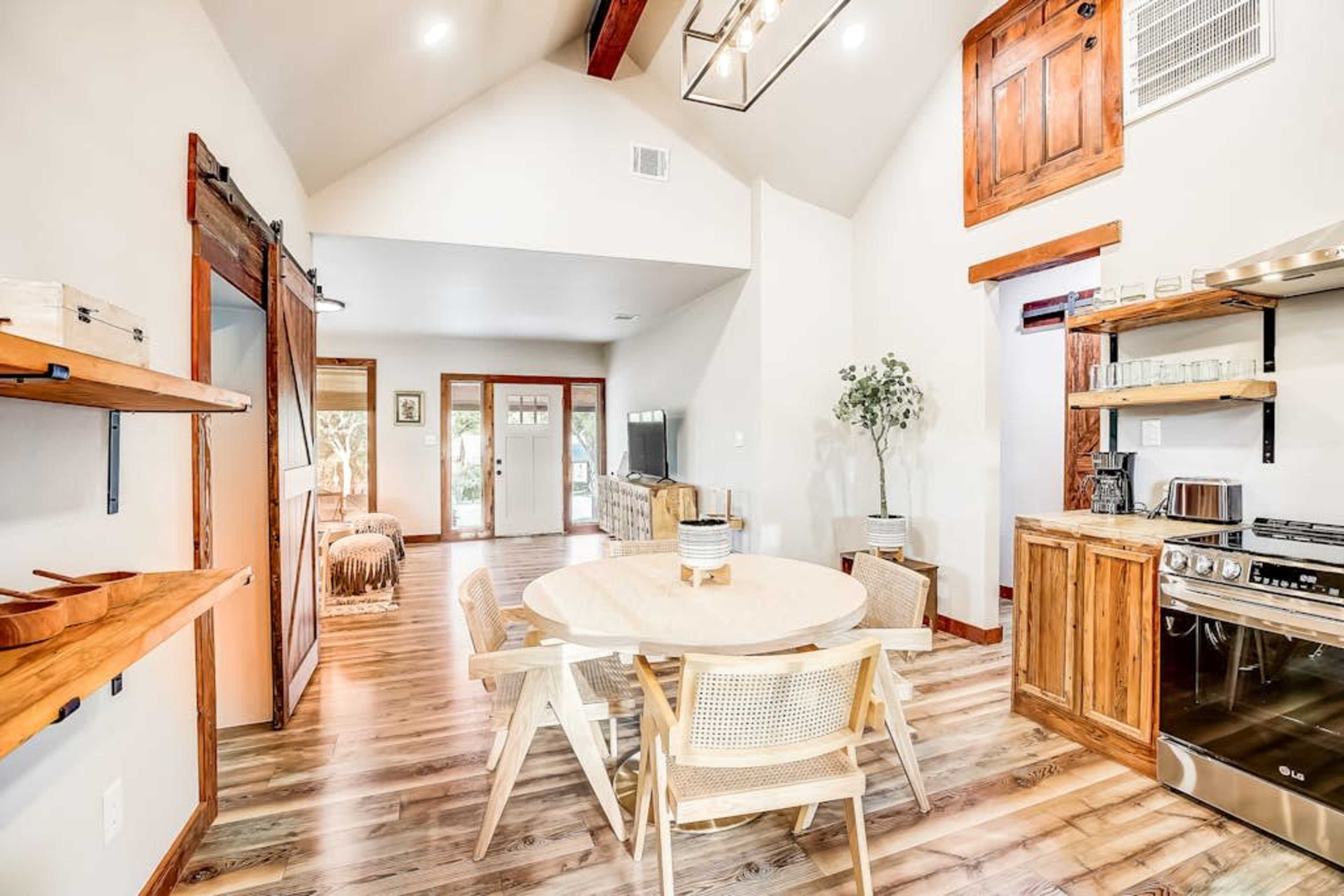 A modern open-concept living space features a round dining table surrounded by light wooden chairs, a rustic kitchen, and large windows allowing natural light to flow in.