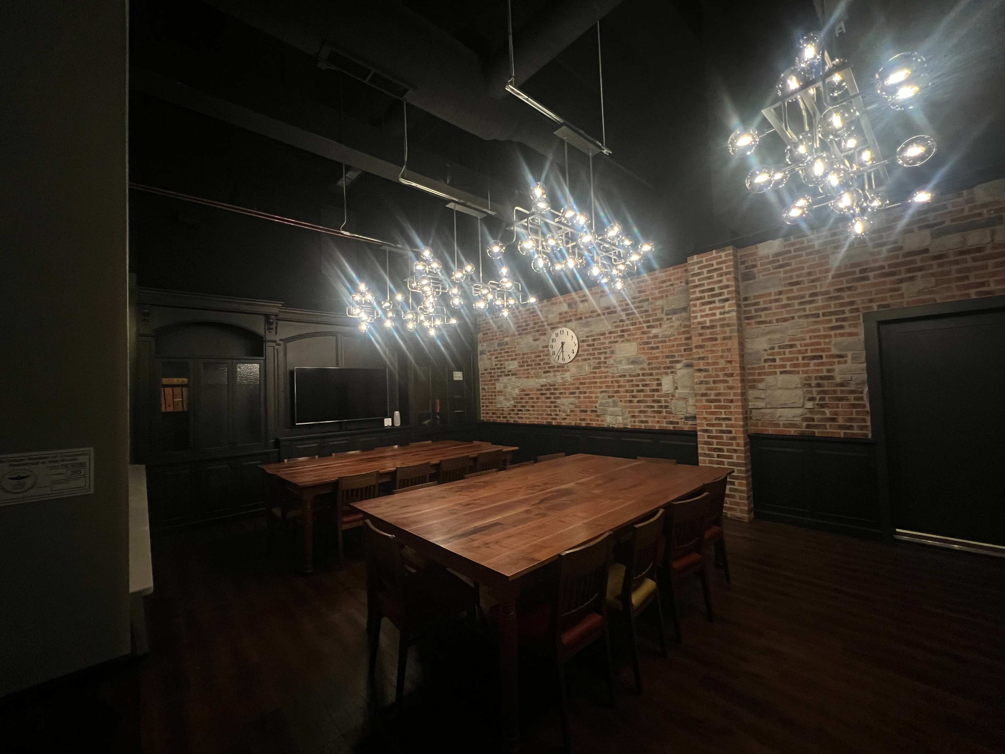 The image shows a large meeting room with a wooden table surrounded by chairs, illuminated by modern chandeliers and featuring exposed brick walls.