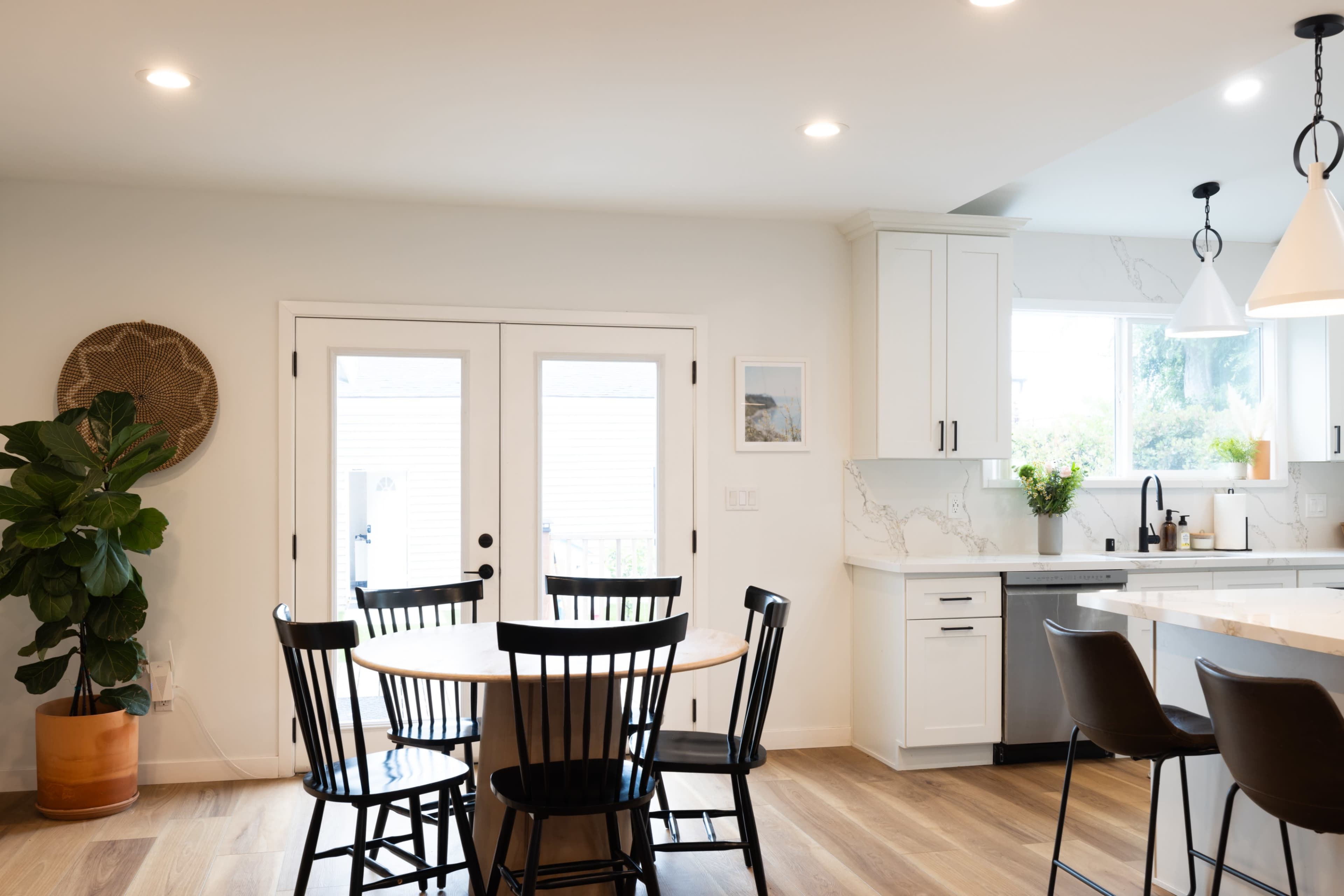 A modern kitchen features a round dining table surrounded by black chairs and opens to a light-filled area with double doors.