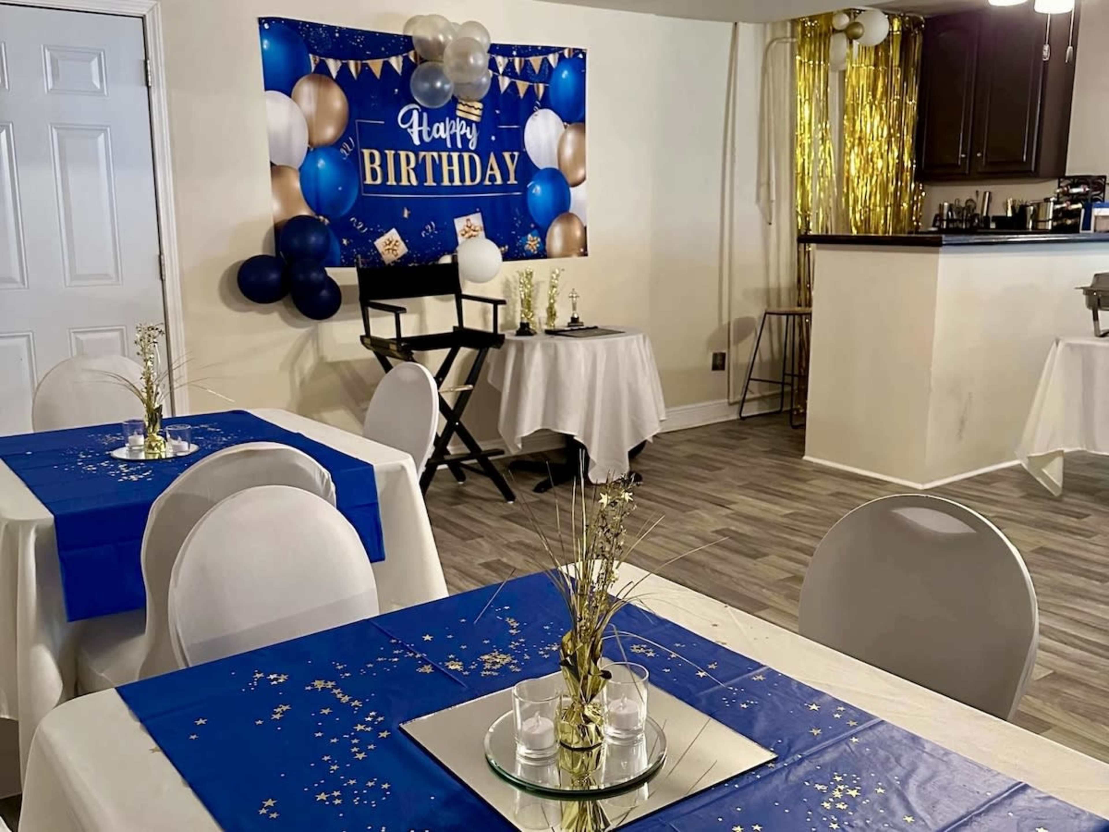 The image shows a birthday party setup with blue and white decorations, tables covered in blue tablecloths, and a wall banner that reads "Happy Birthday."