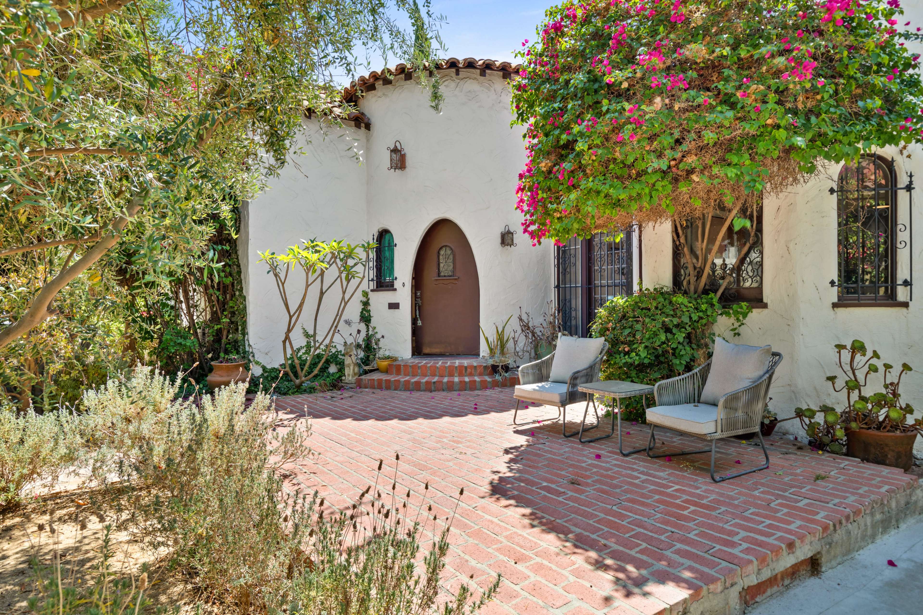 A small, white stucco house with a rounded wooden door is surrounded by a brick patio and flowering plants.