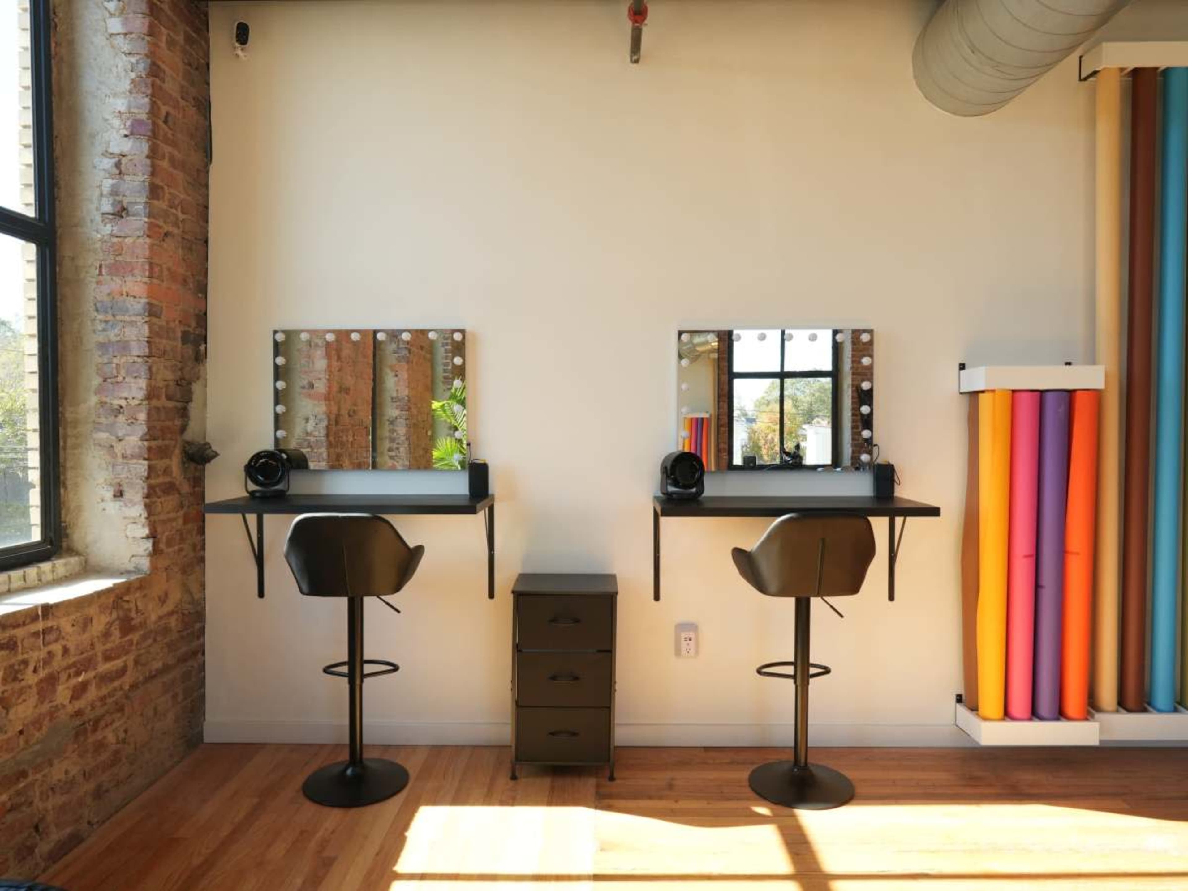 The image shows a brightly lit room with two makeup stations featuring mirrors and studio lights, alongside a small storage unit and rolls of colorful backdrop paper.