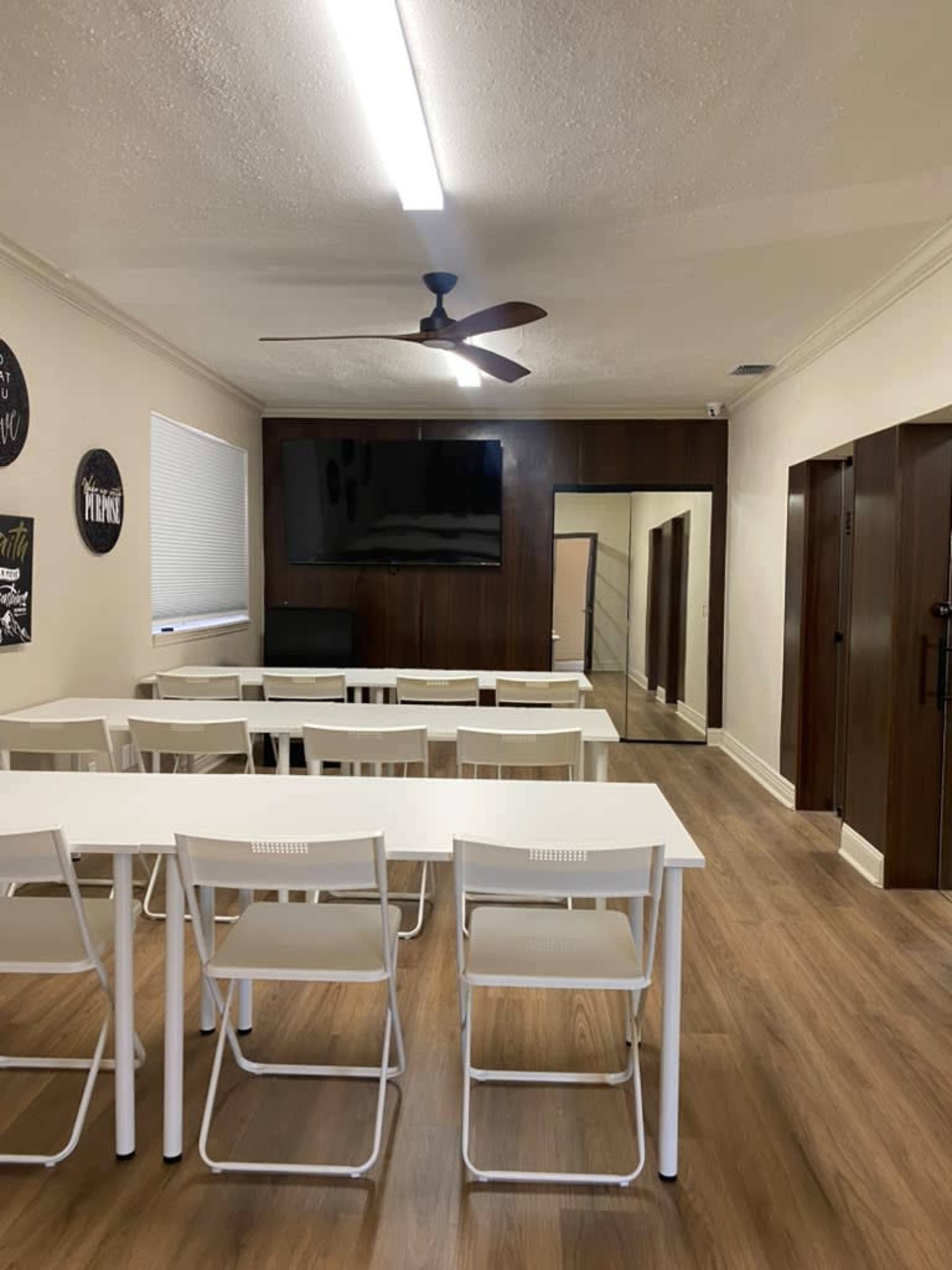 The room features a long table with white chairs, a large flat-screen TV mounted on the wall, and wood-paneled storage areas along with a mirror in the background.