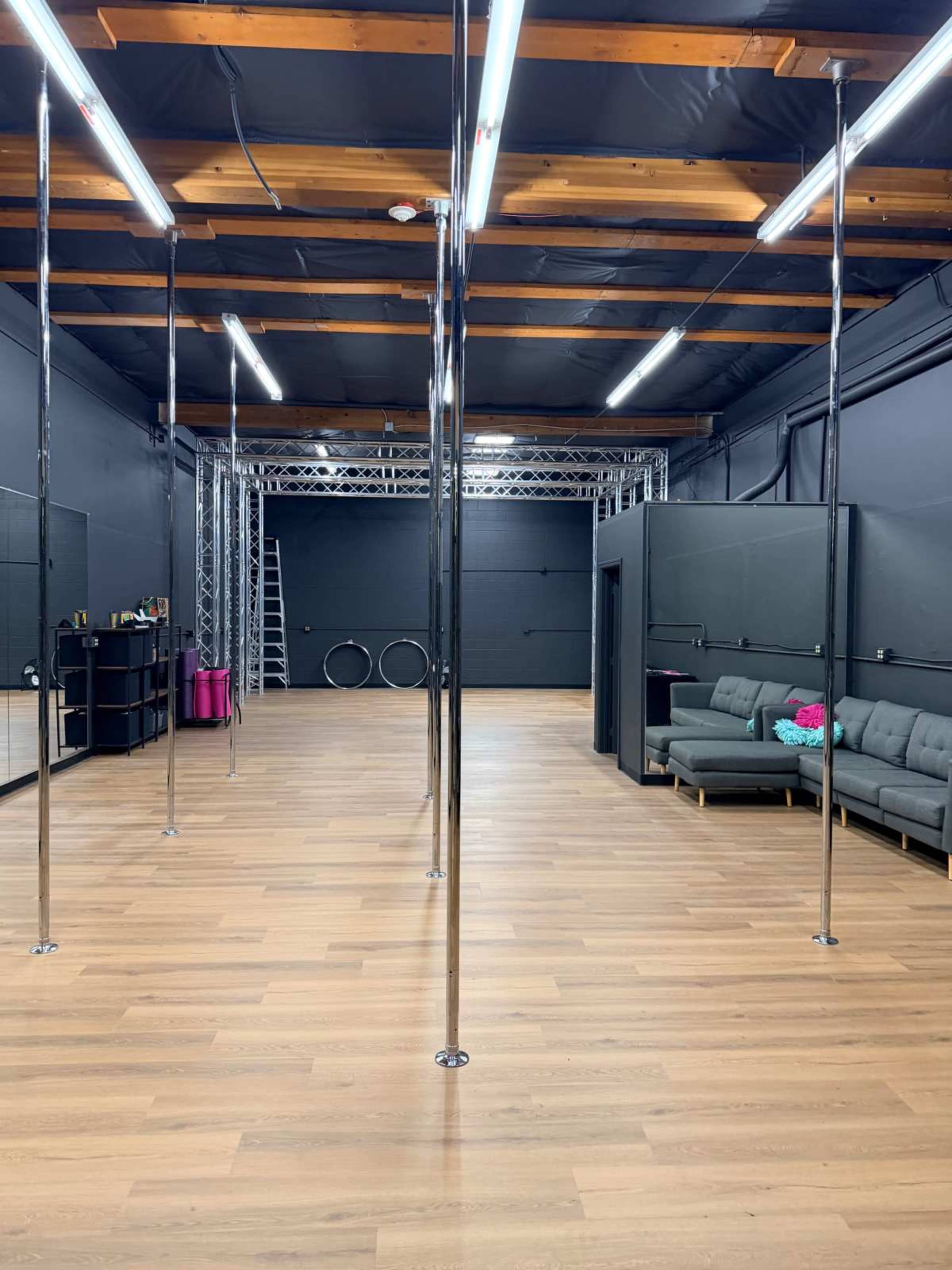 The image shows an empty dance studio with polished wooden floors, several vertical poles, and a lounge area with gray sofas.