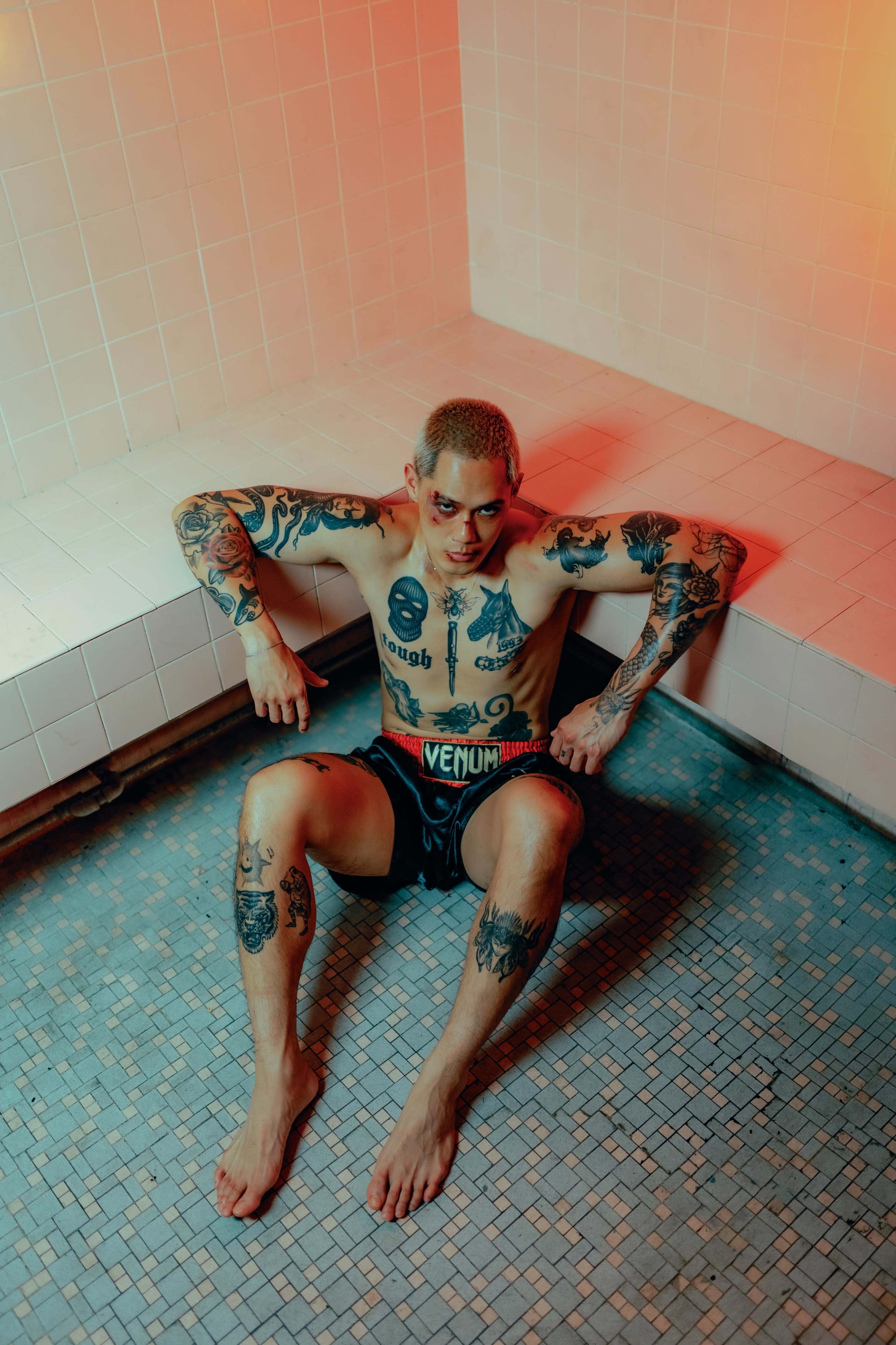 A tattooed individual sits on the edge of a tiled shower area, with an orange light illuminating the scene.