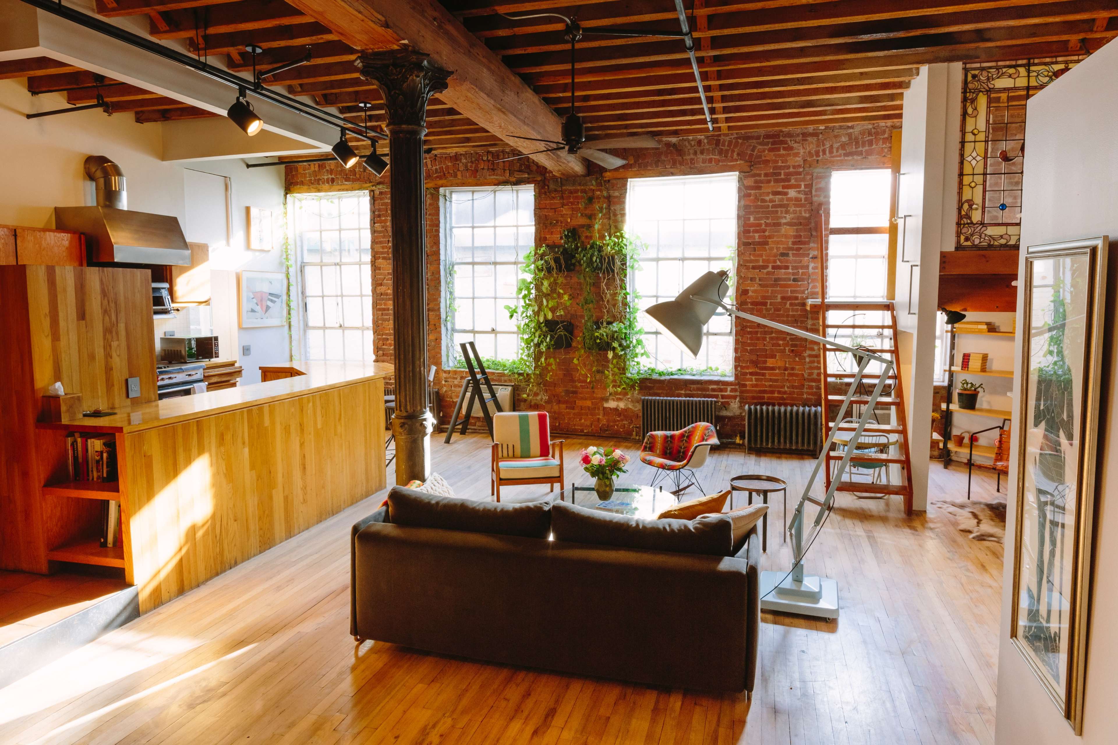 The image shows a spacious loft-style living area featuring exposed brick walls, wooden beams, and modern furnishings, including a couch, a dining area, and various indoor plants.