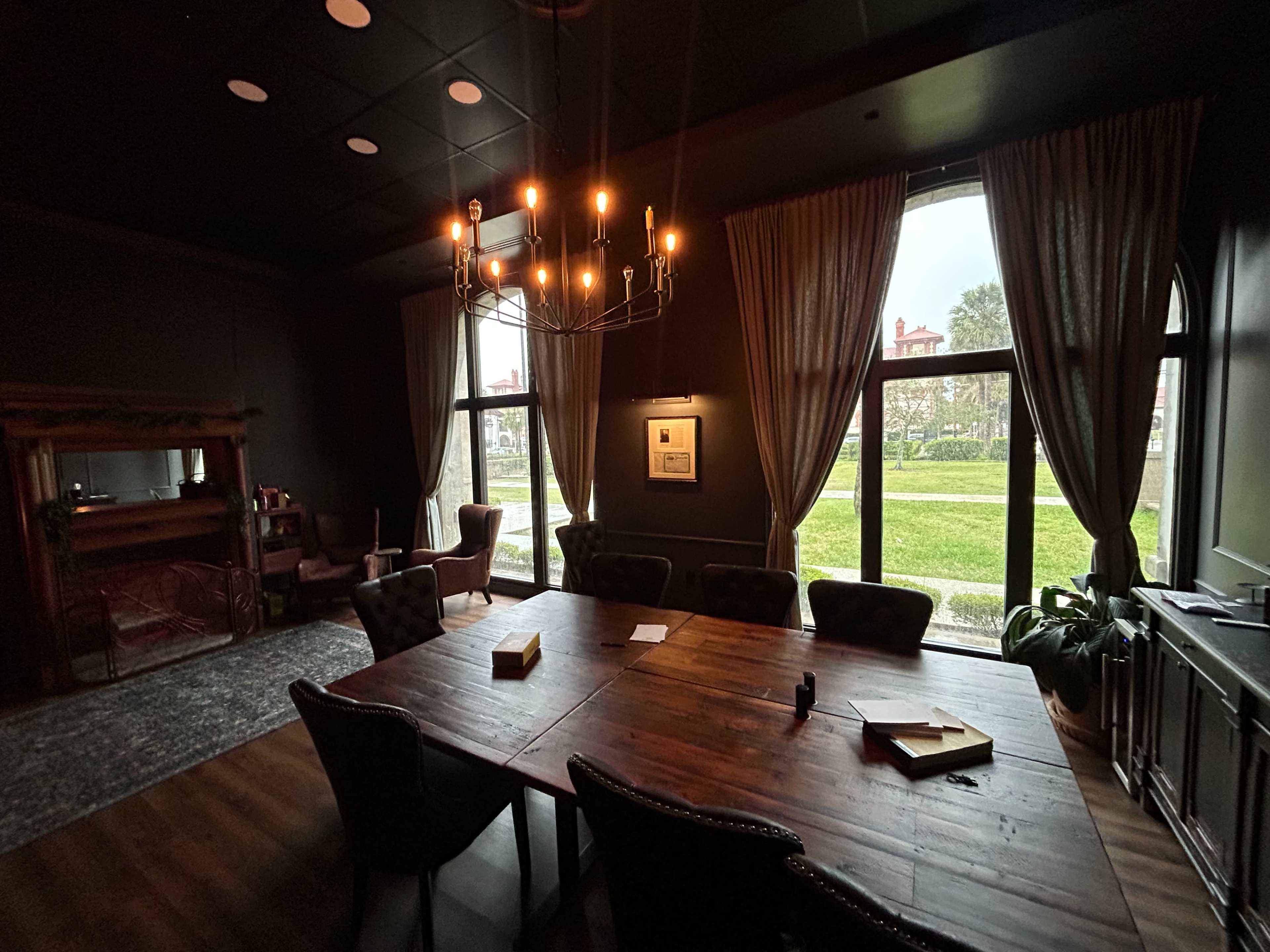 The image shows a dimly lit room with a large wooden table surrounded by chairs, featuring large windows that provide a view of a grassy area outside.