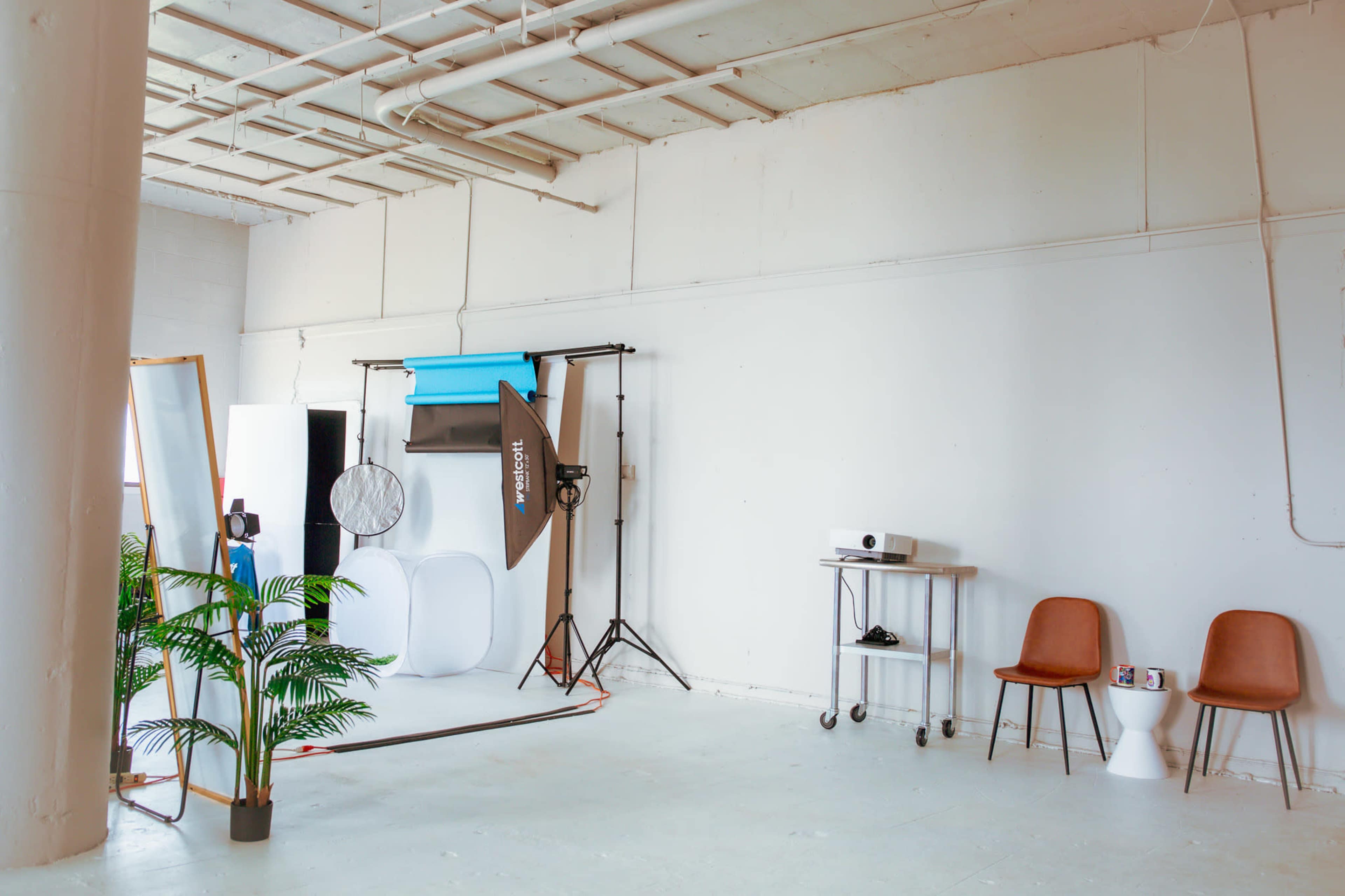 A minimalist photography studio featuring a white backdrop, lighting equipment, a projector, and various furniture pieces.