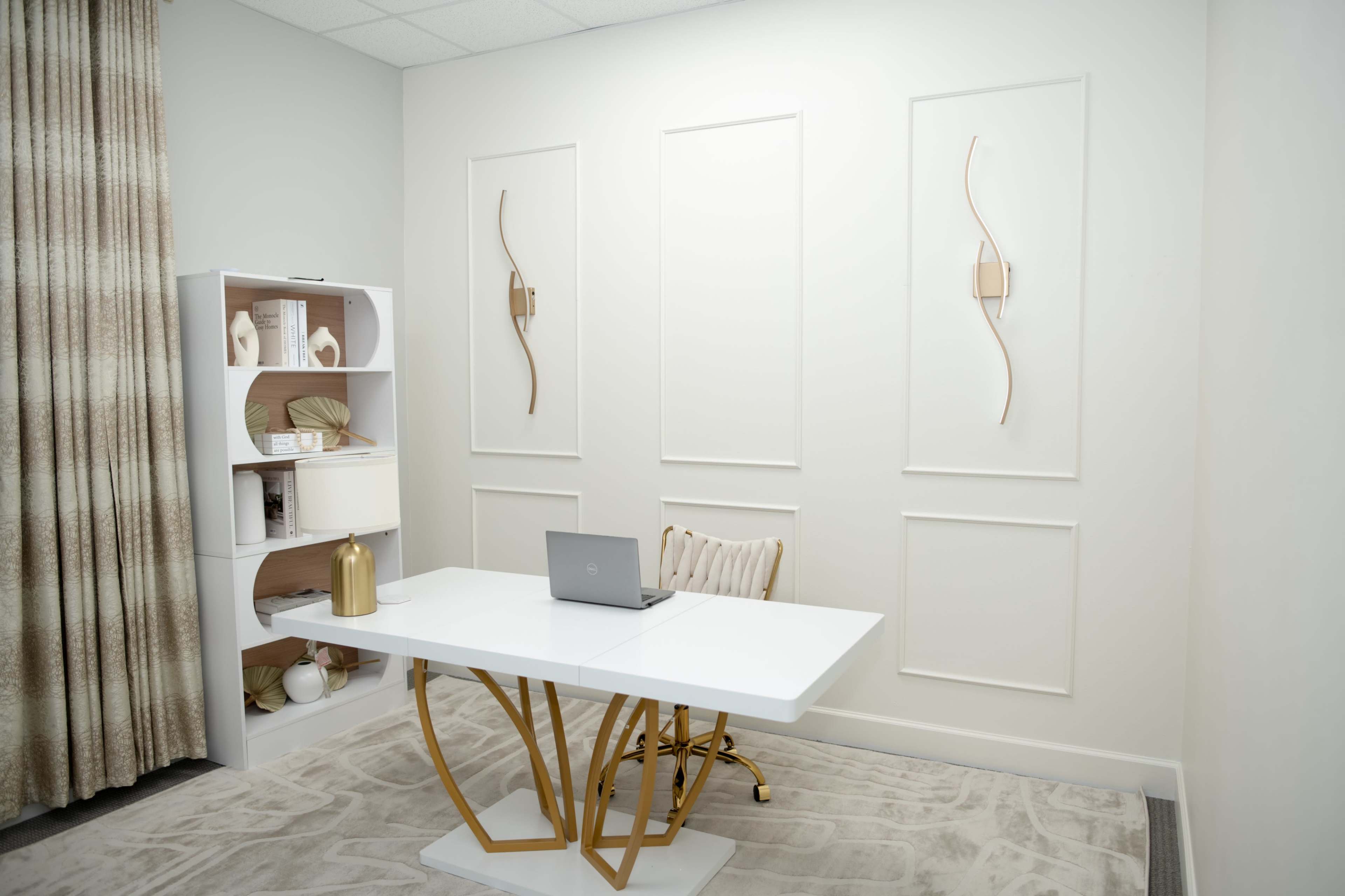 The image shows a modern office space with a white desk and a laptop, accompanied by a decorative bookshelf and neutral-colored curtains.