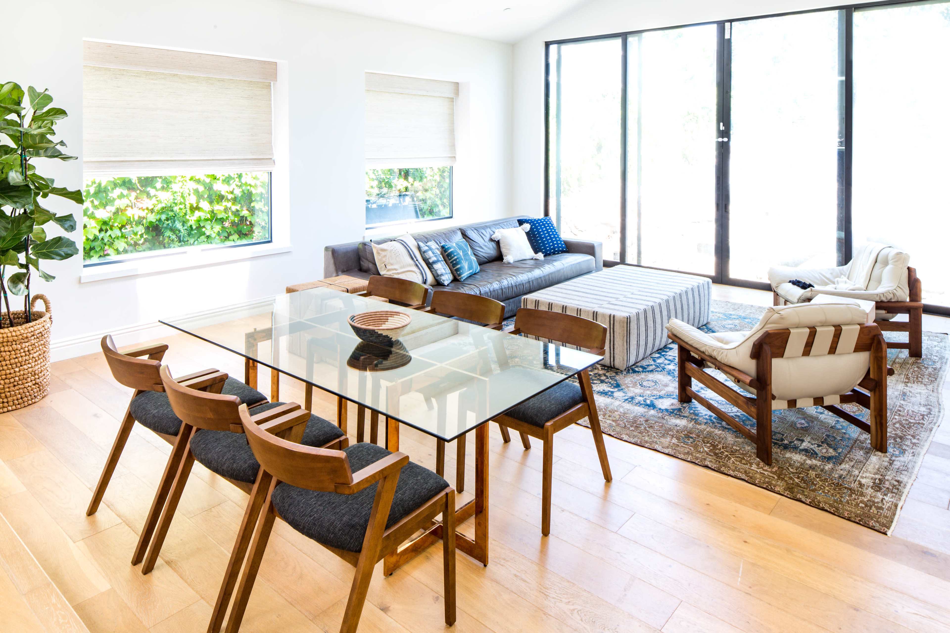 A modern living and dining area features a glass-top table with wooden chairs, a sectional sofa, and large windows providing natural light.