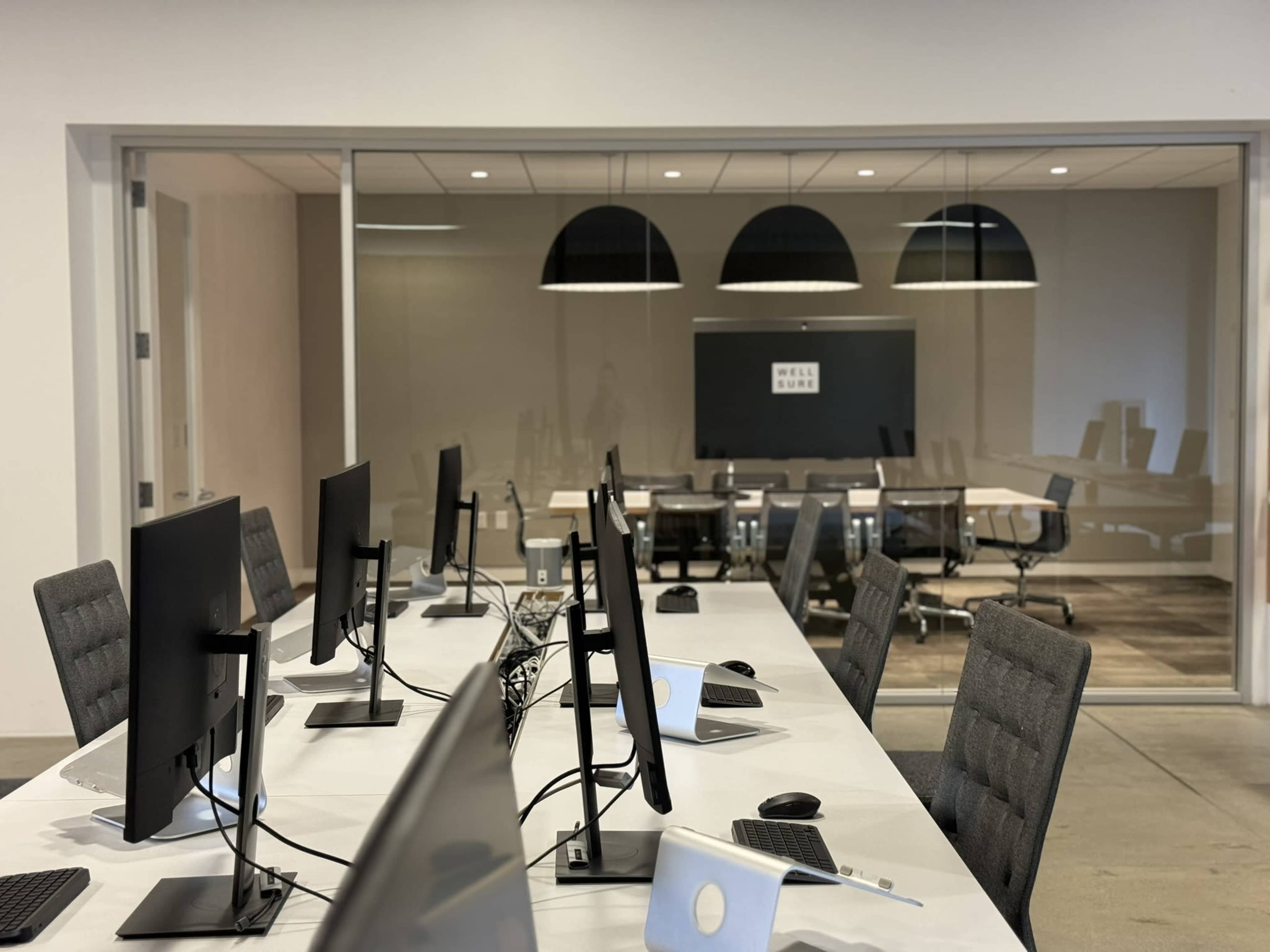 A modern office space with multiple computer workstations facing an adjacent conference room featuring a long table and several chairs.