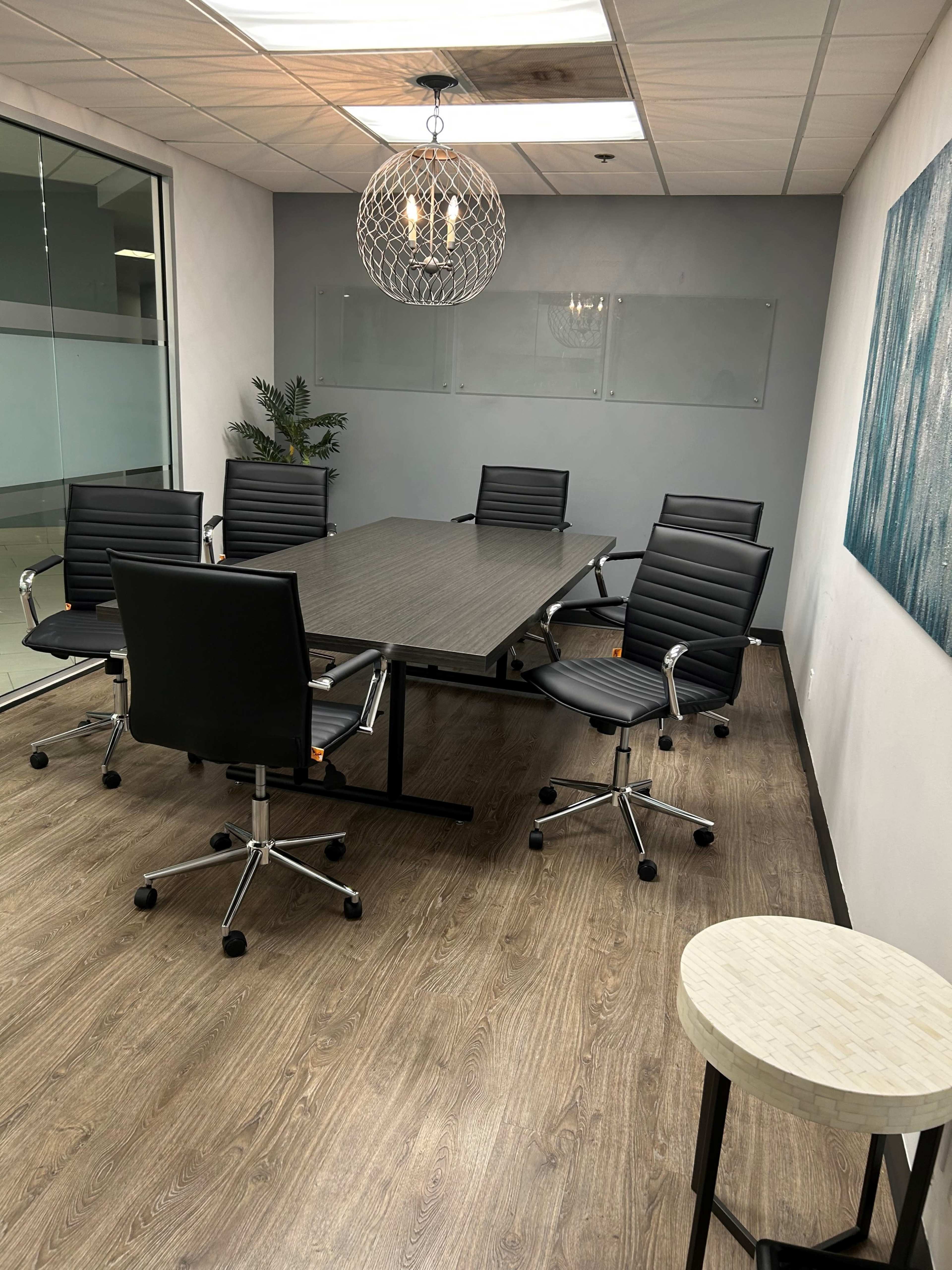 The image shows a modern conference room featuring a large rectangular table surrounded by six black office chairs, with a decorative light fixture overhead.