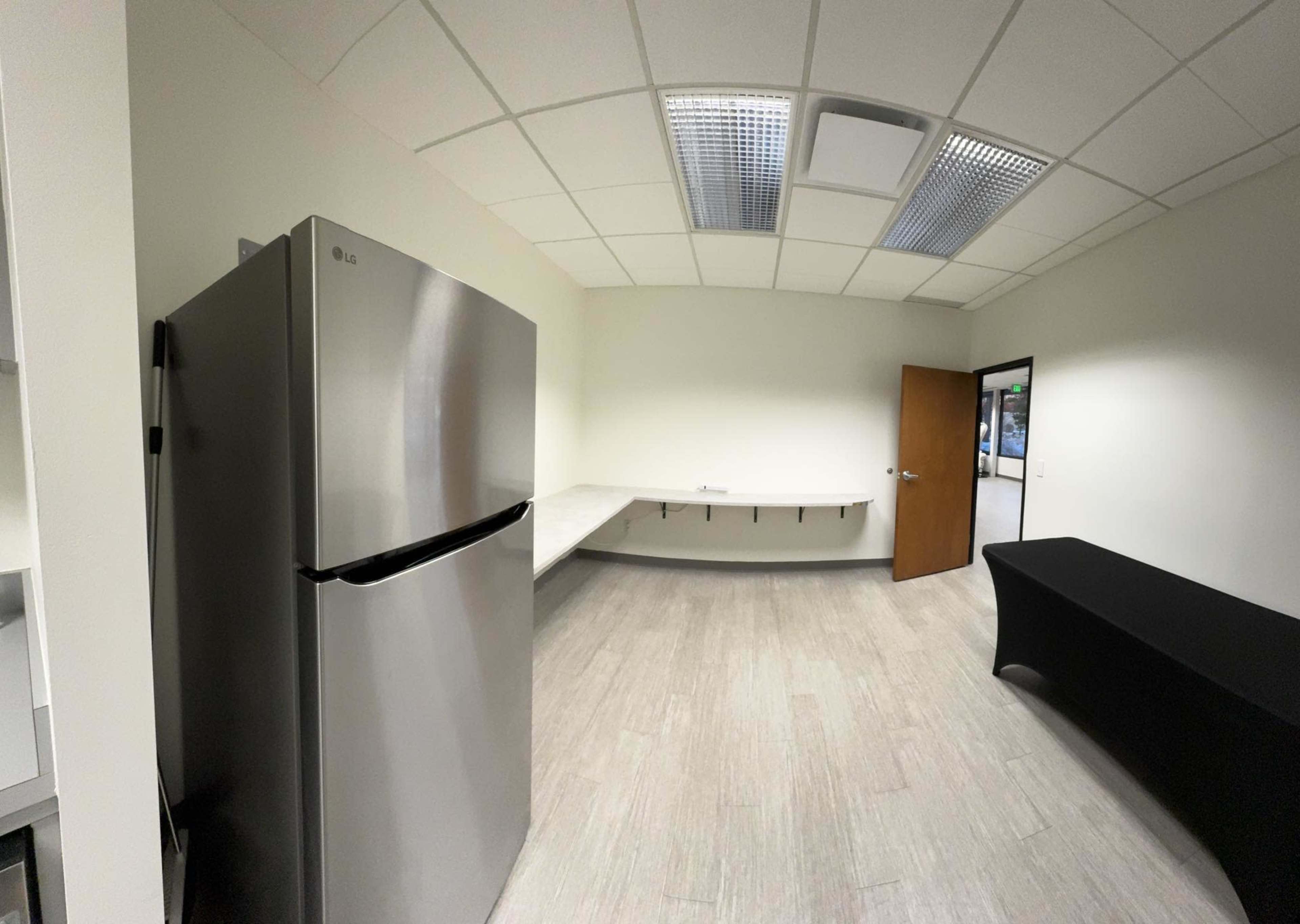 The image shows a bright, empty room featuring a stainless steel refrigerator, a long counter, and a black table against one wall.