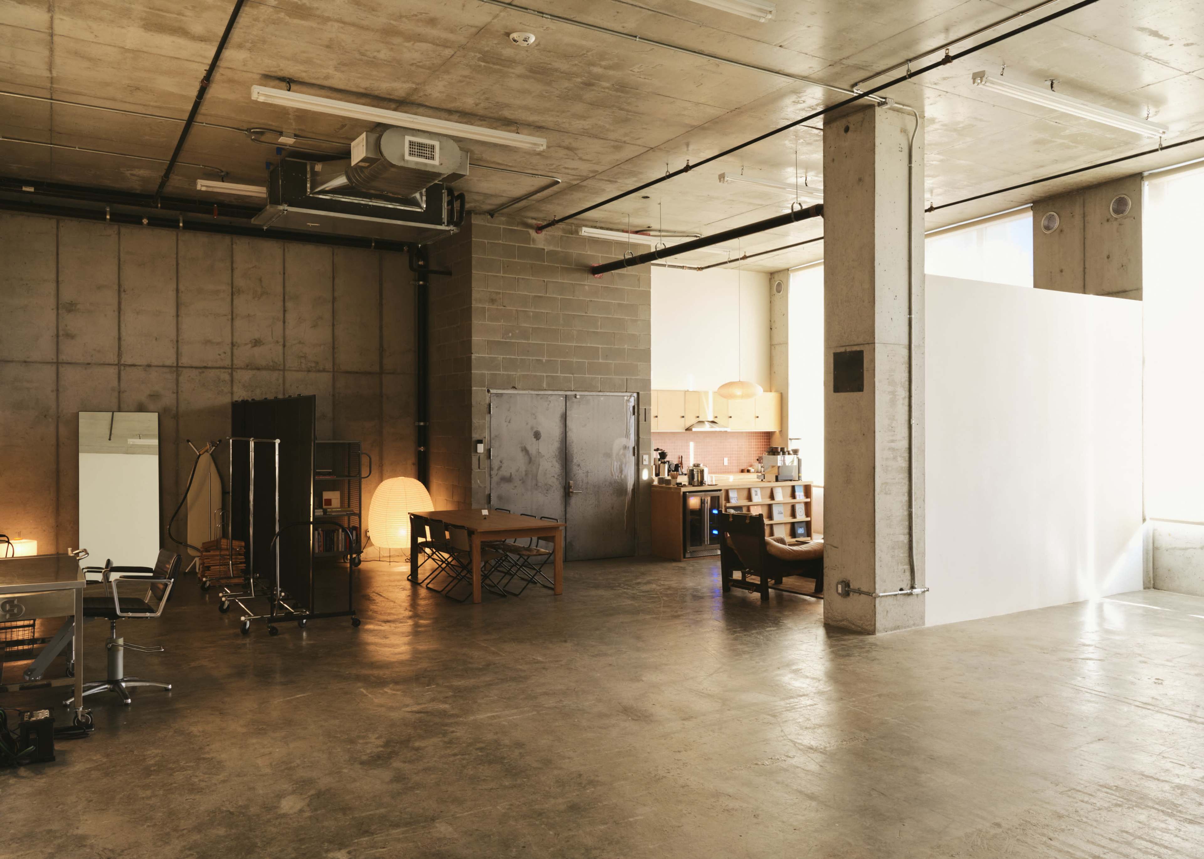 The image shows a spacious industrial-style room with concrete walls, a minimalist kitchen area, a large window, and various furniture pieces including tables and chairs.