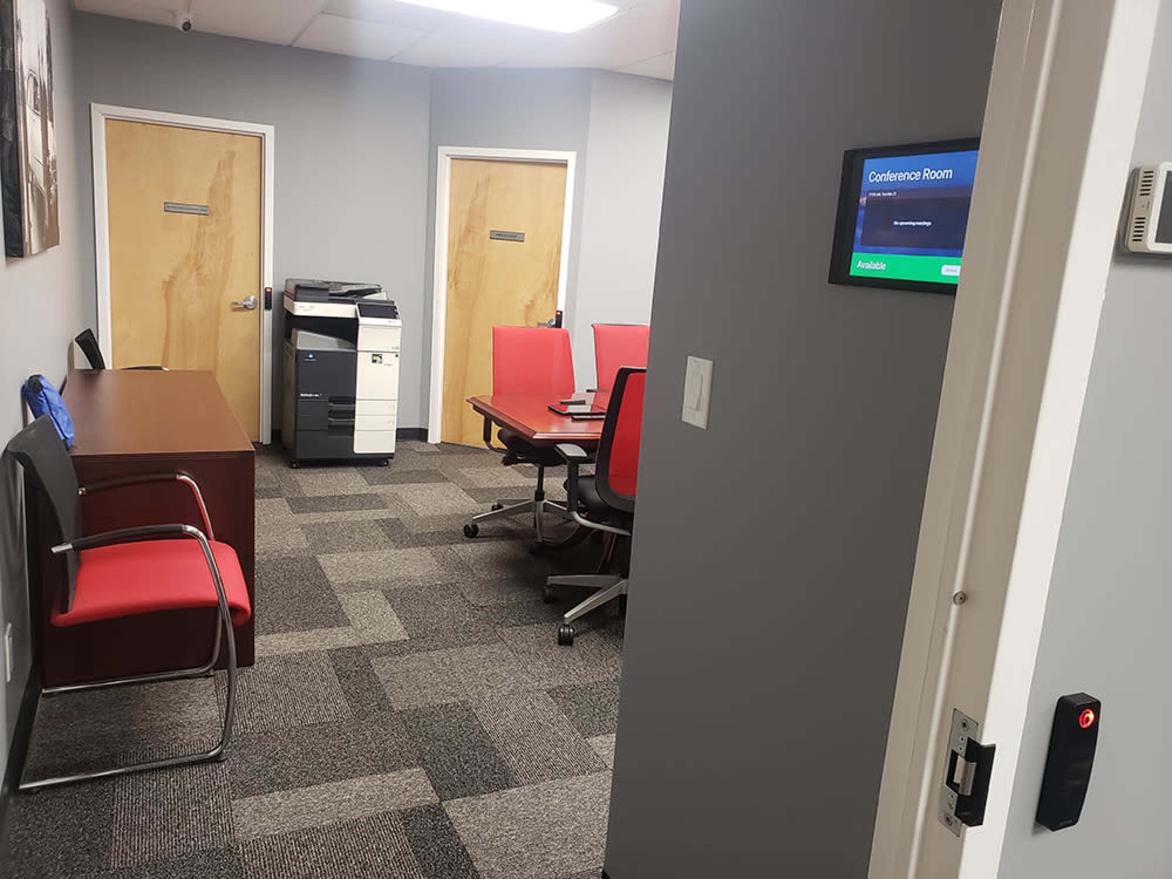 A conference room with red chairs, a table, and a printer, viewed from an office doorway.