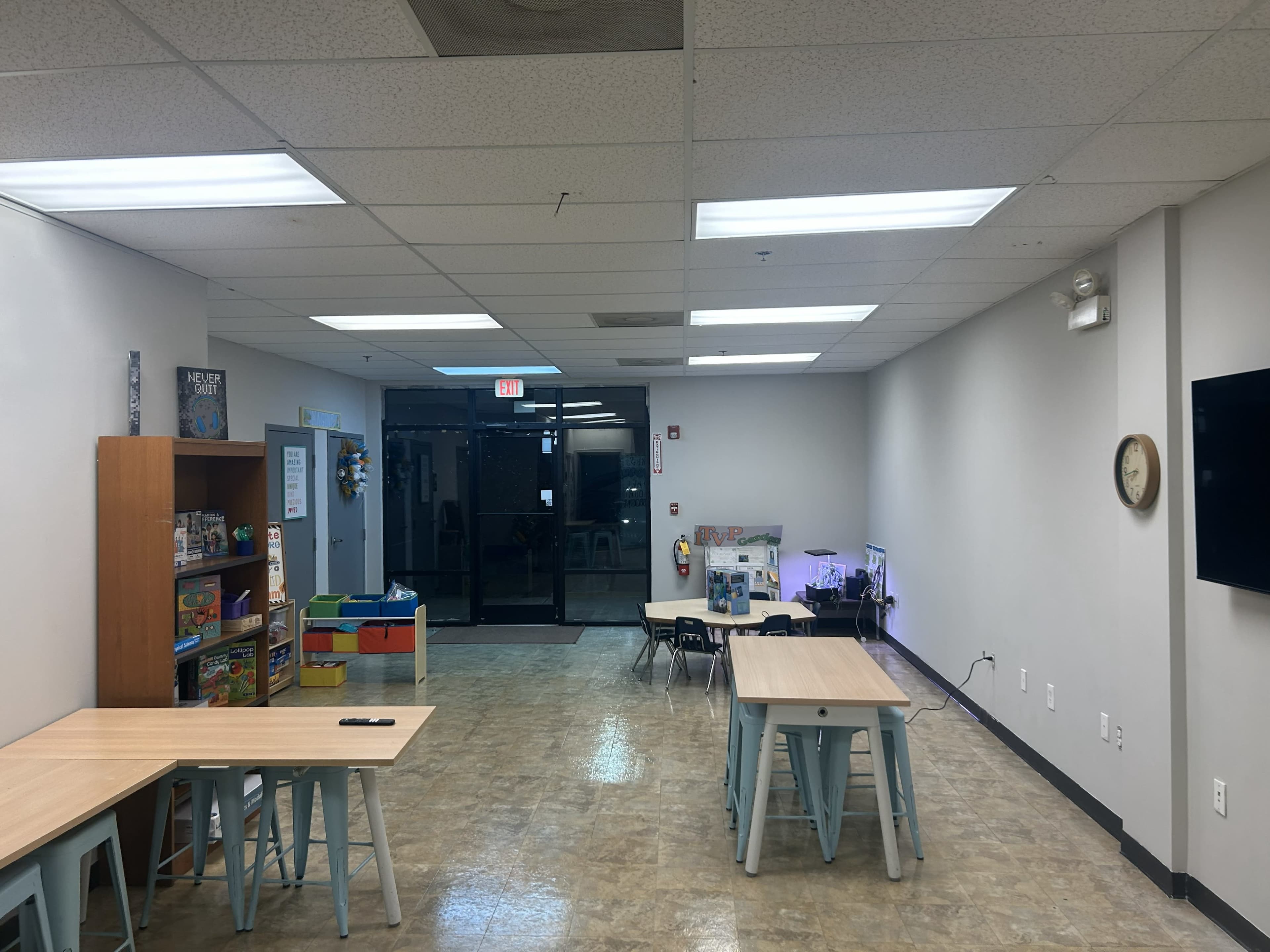 The image shows a bright, spacious classroom with several tables, a bookshelf filled with educational materials, and an entrance visible in the background.