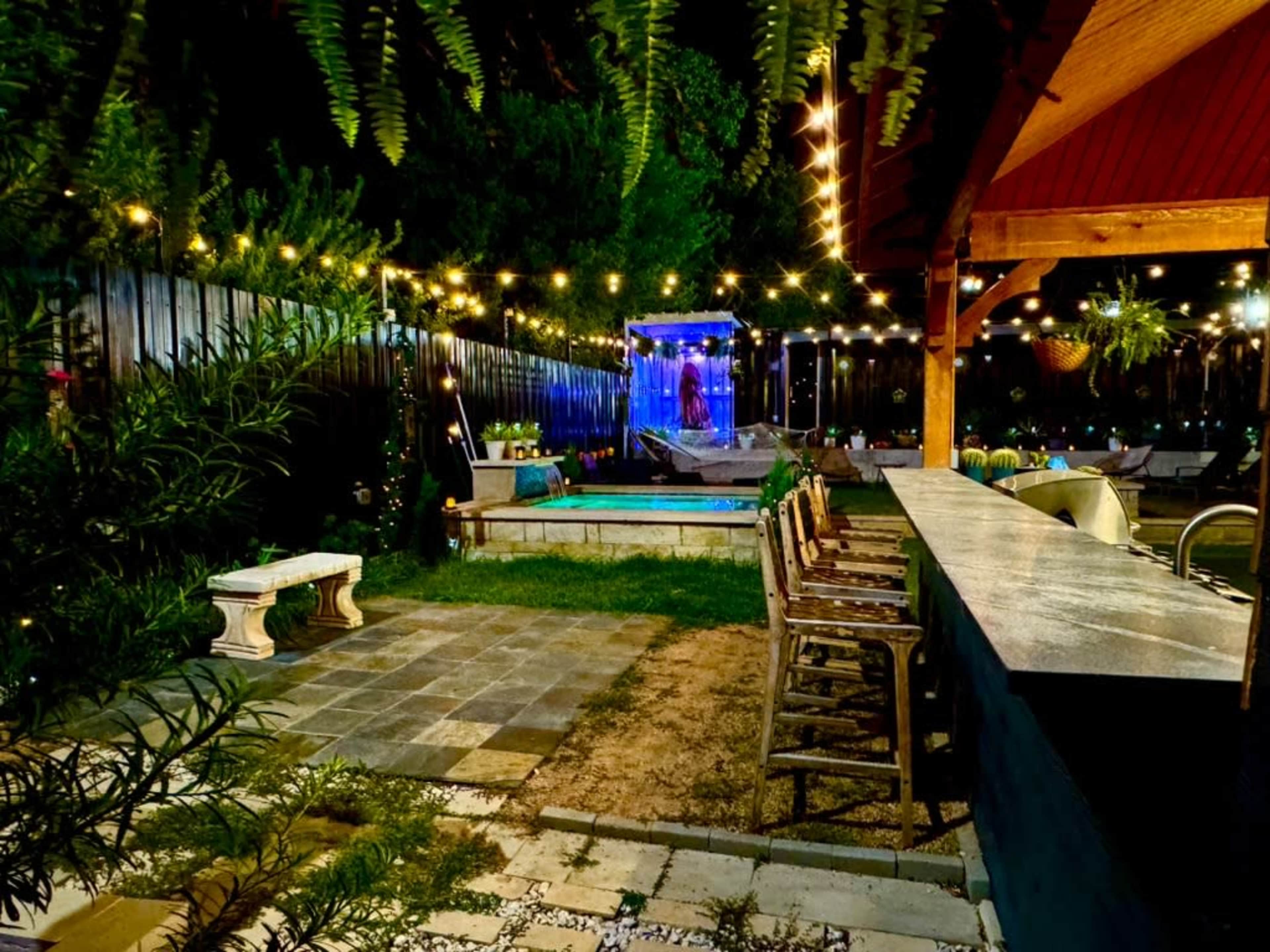 The image shows a backyard patio with a bar area, a small pool, and string lights illuminating the space at night.