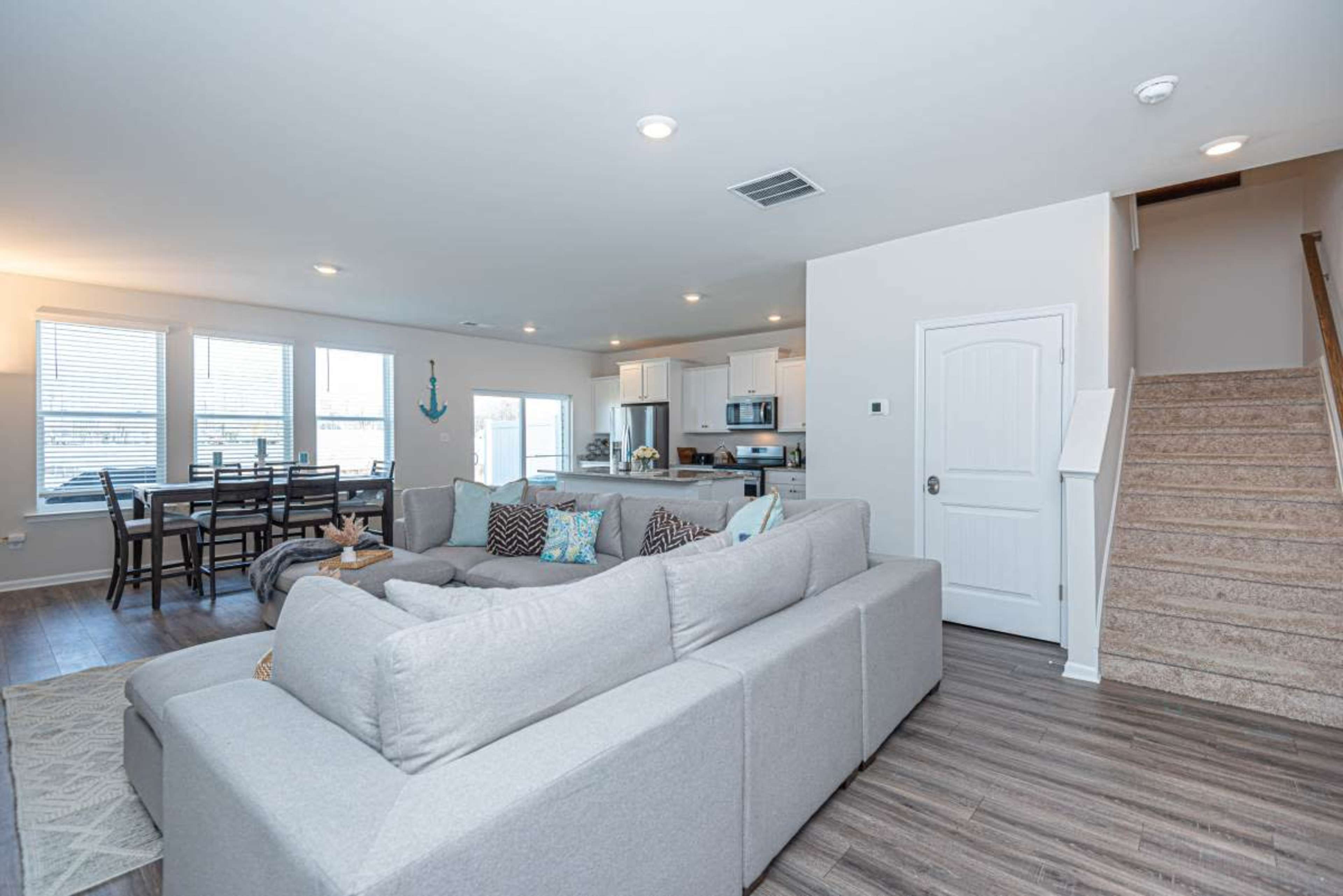 A modern living room with a large sectional sofa, a dining area with a table and chairs, and a staircase leading to an upper level.