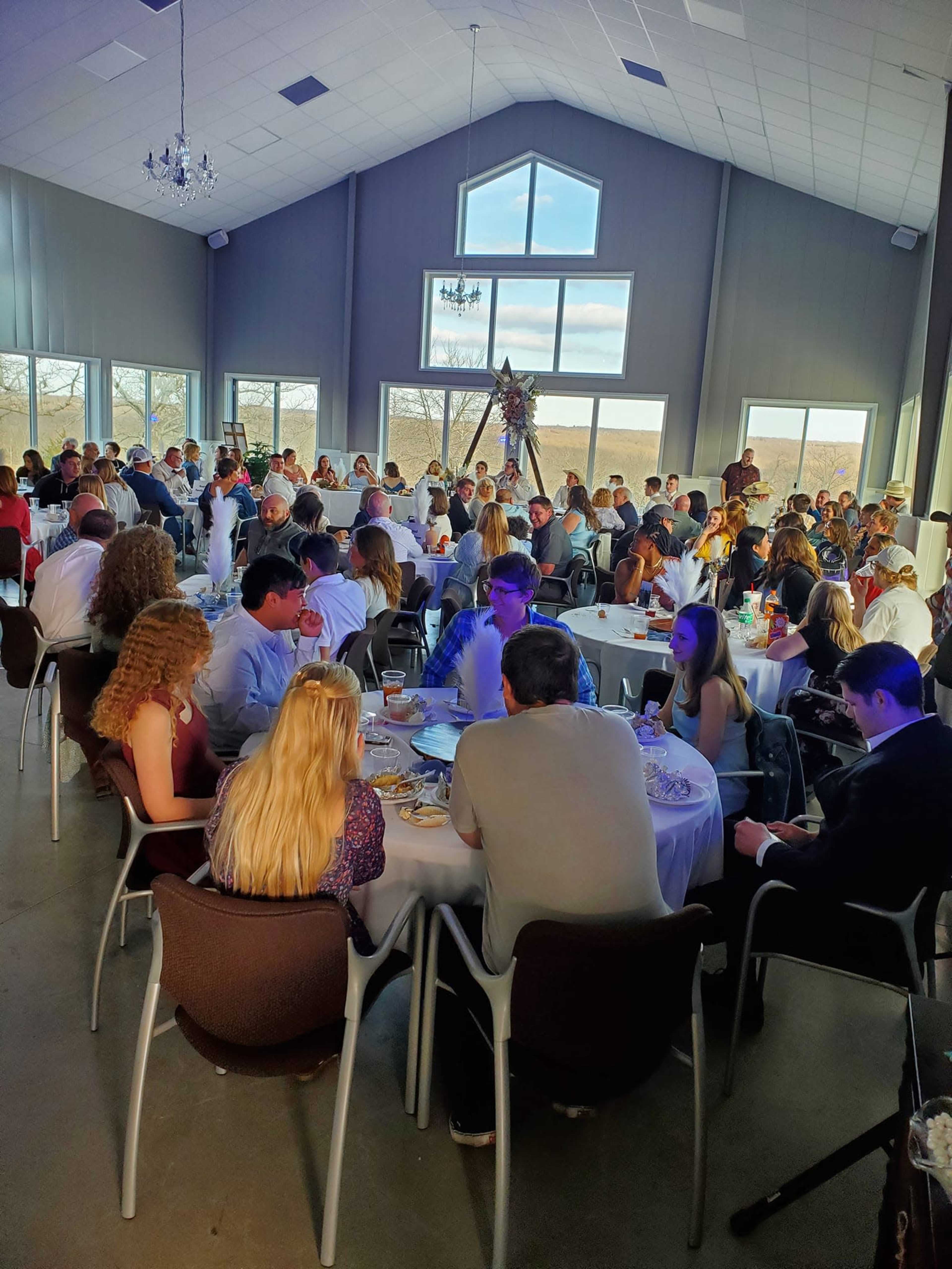 A large gathering is taking place in a well-lit indoor event space with tables arranged for dining.
