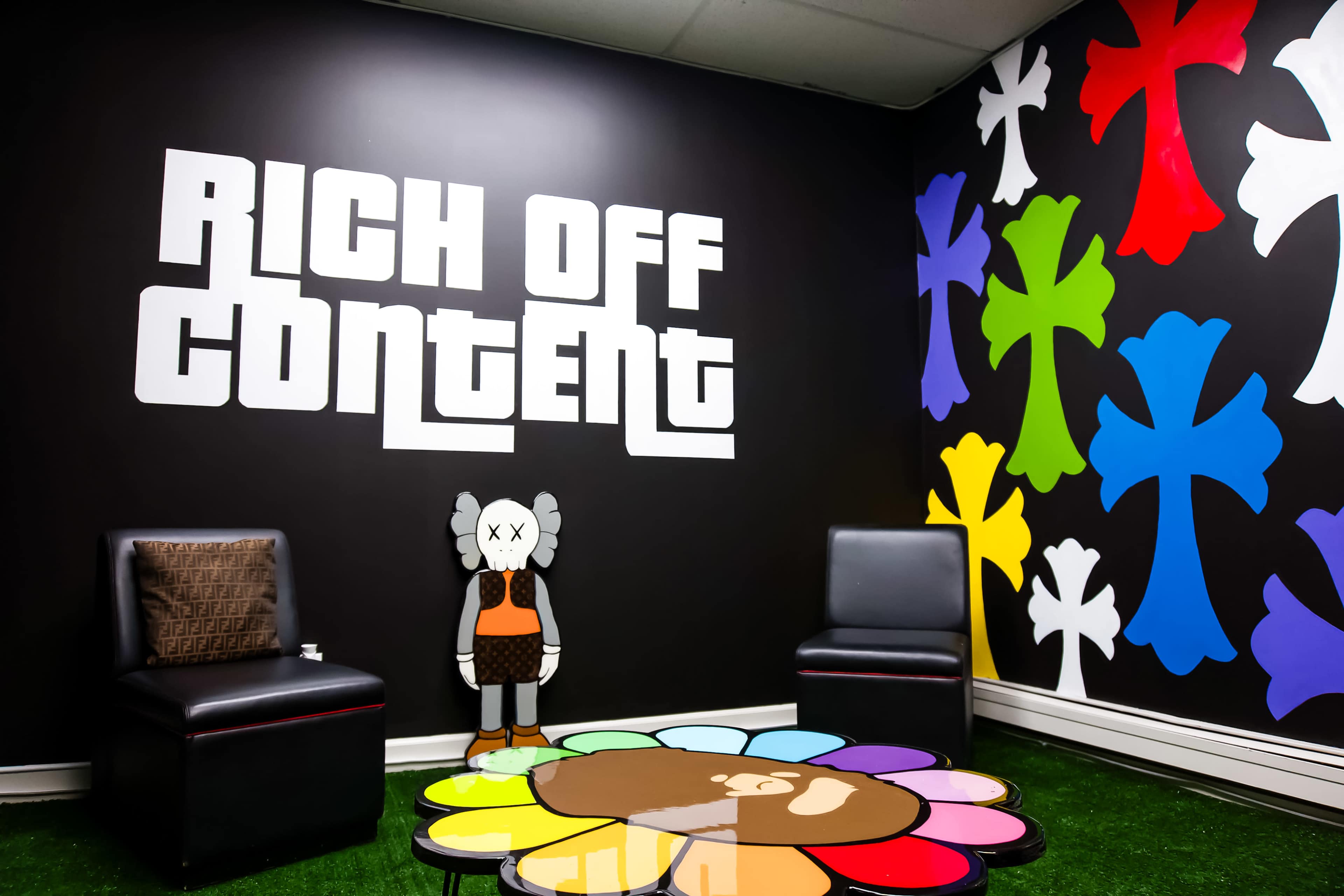 The image shows a modern room featuring a black wall with the phrase "RICH OFF CONTENT" in large white letters, colorful abstract designs in various shapes on other walls, and a decorative character beside a flower-shaped table.