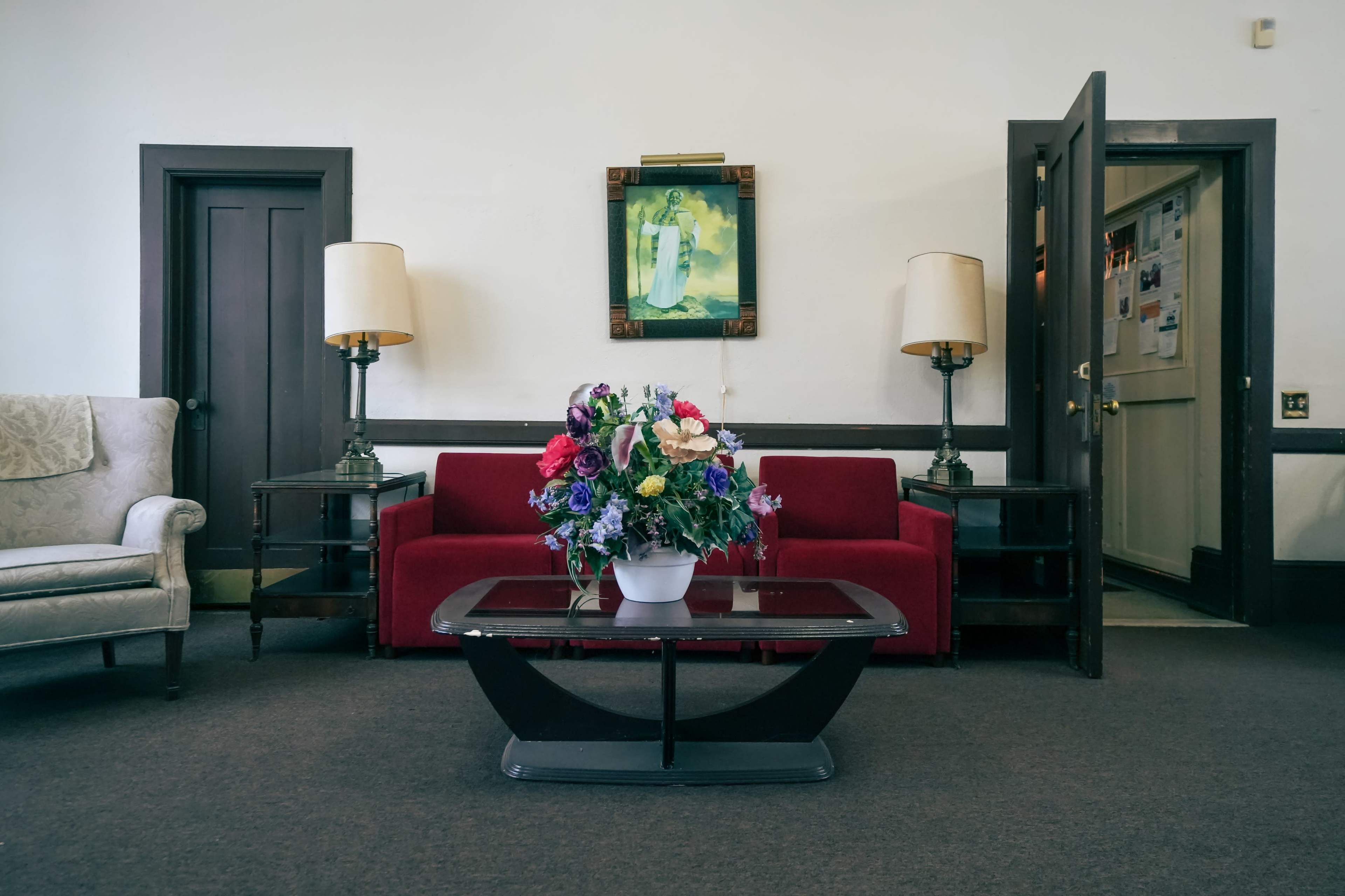 A living room features a red sofa, a coffee table with a vase of flowers, and two lamps beside a beige armchair, with a door leading to another room in the background.