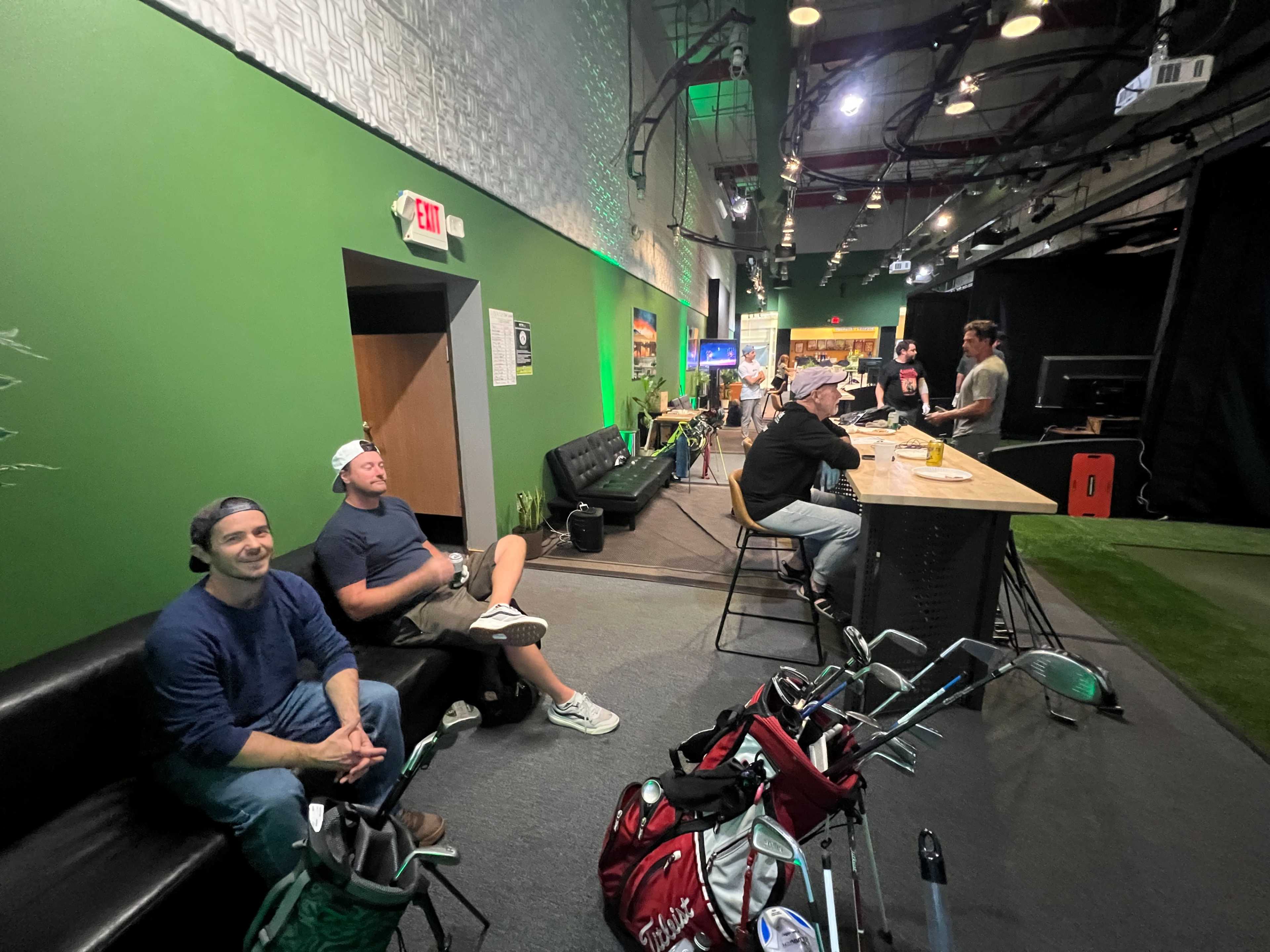 A group of people is seated at a bar with golf clubs nearby, while another person stands at a counter in a green-walled indoor golf facility.