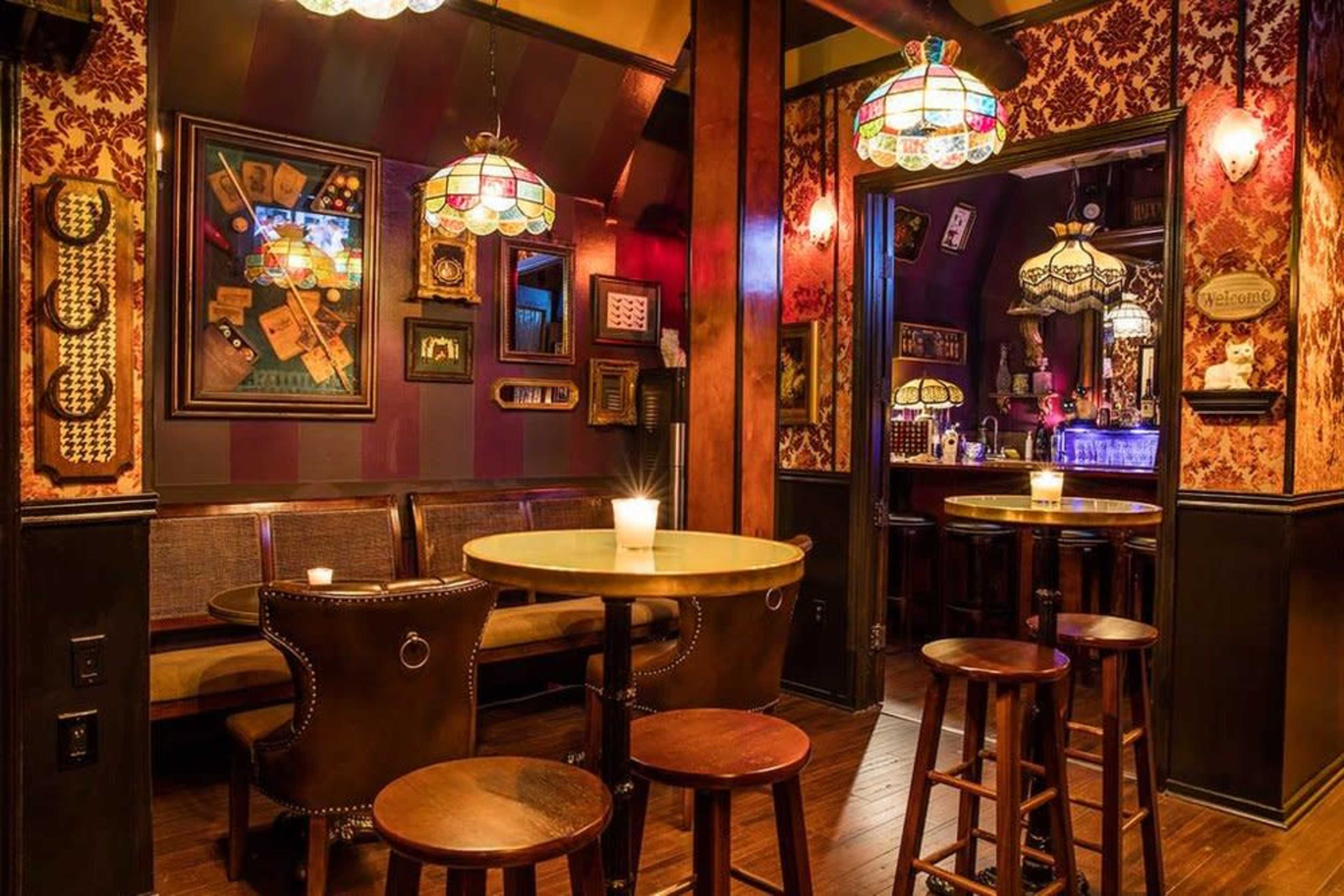 A dimly lit bar interior with stained glass lamps, ornate wallpaper, and wooden furnishings including a round table and high stools.