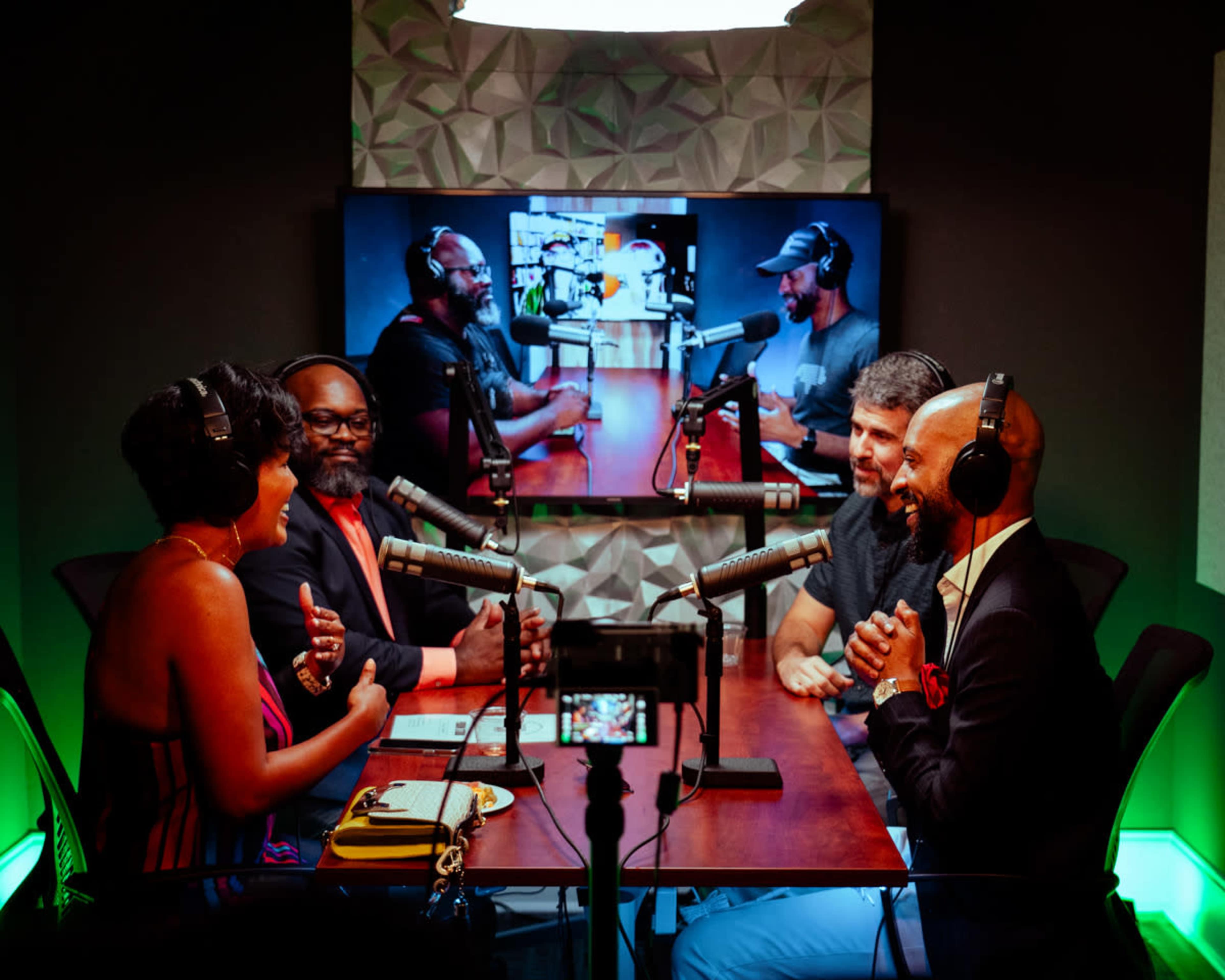 A group of four people engages in a discussion at a podcast recording studio, with a large screen displaying their conversation in the background.
