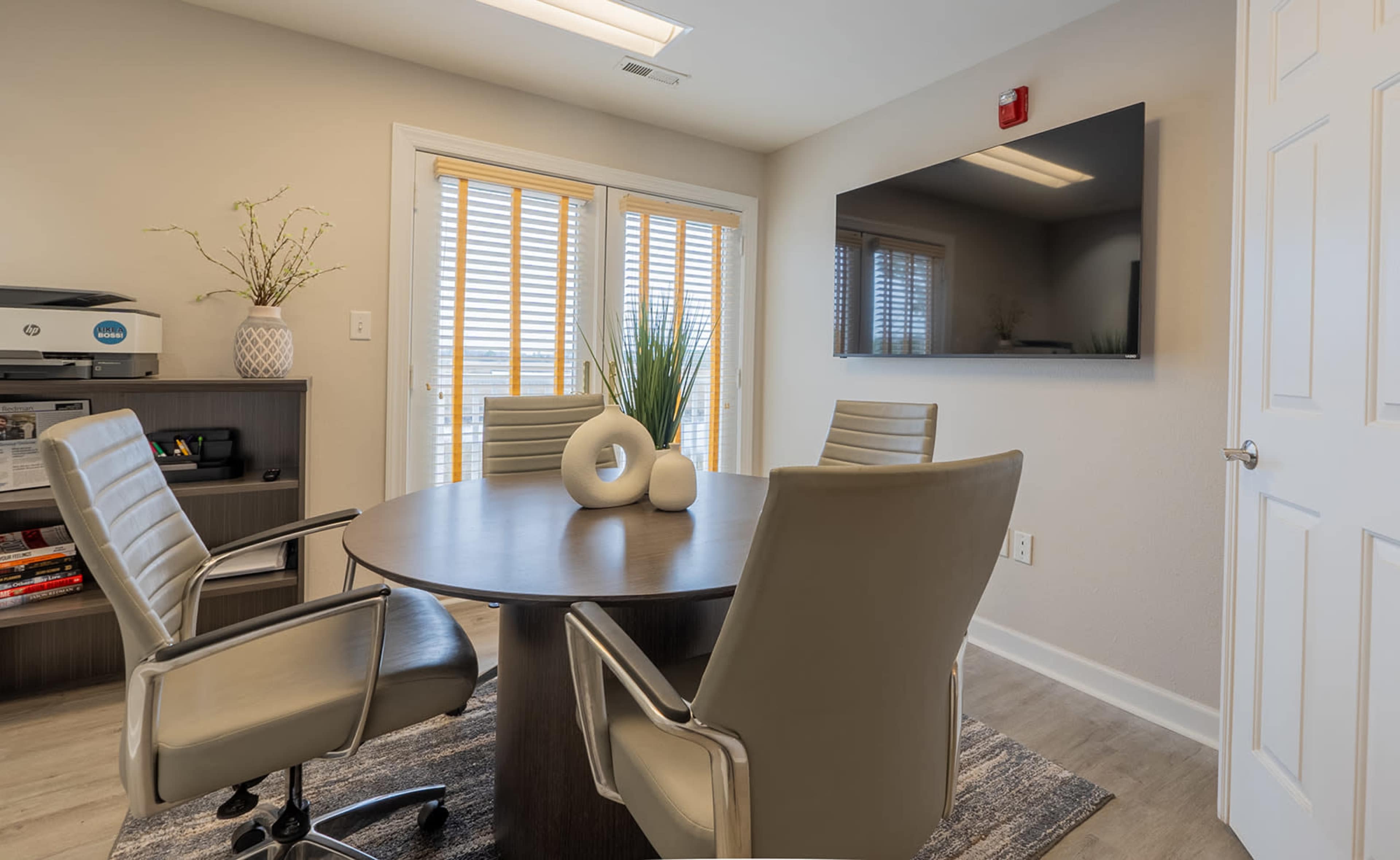 A modern meeting room featuring a round table surrounded by four chairs, with a large wall-mounted TV and window blinds in the background.