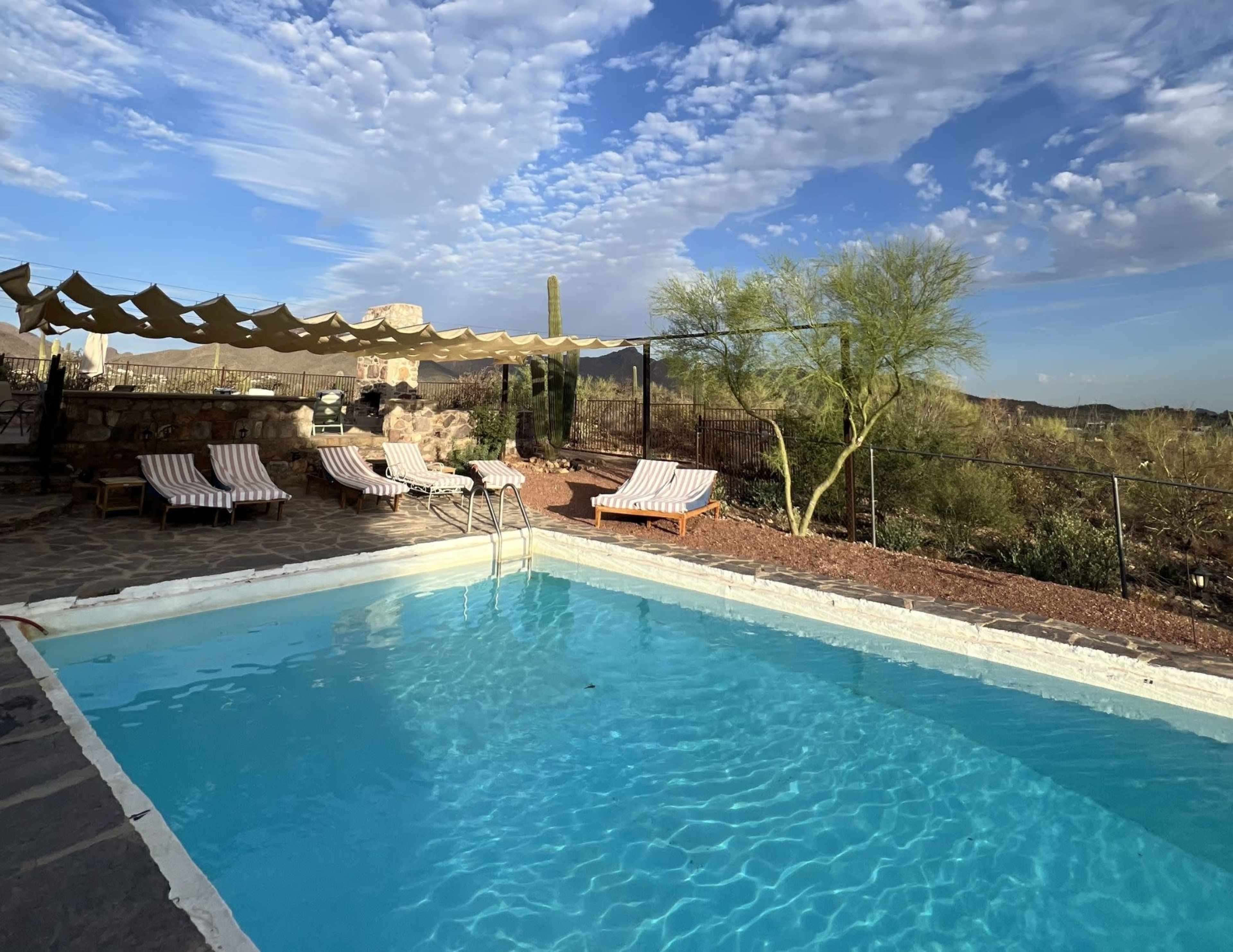 The image shows a tranquil outdoor swimming pool surrounded by lounge chairs and desert vegetation under a partly cloudy sky.