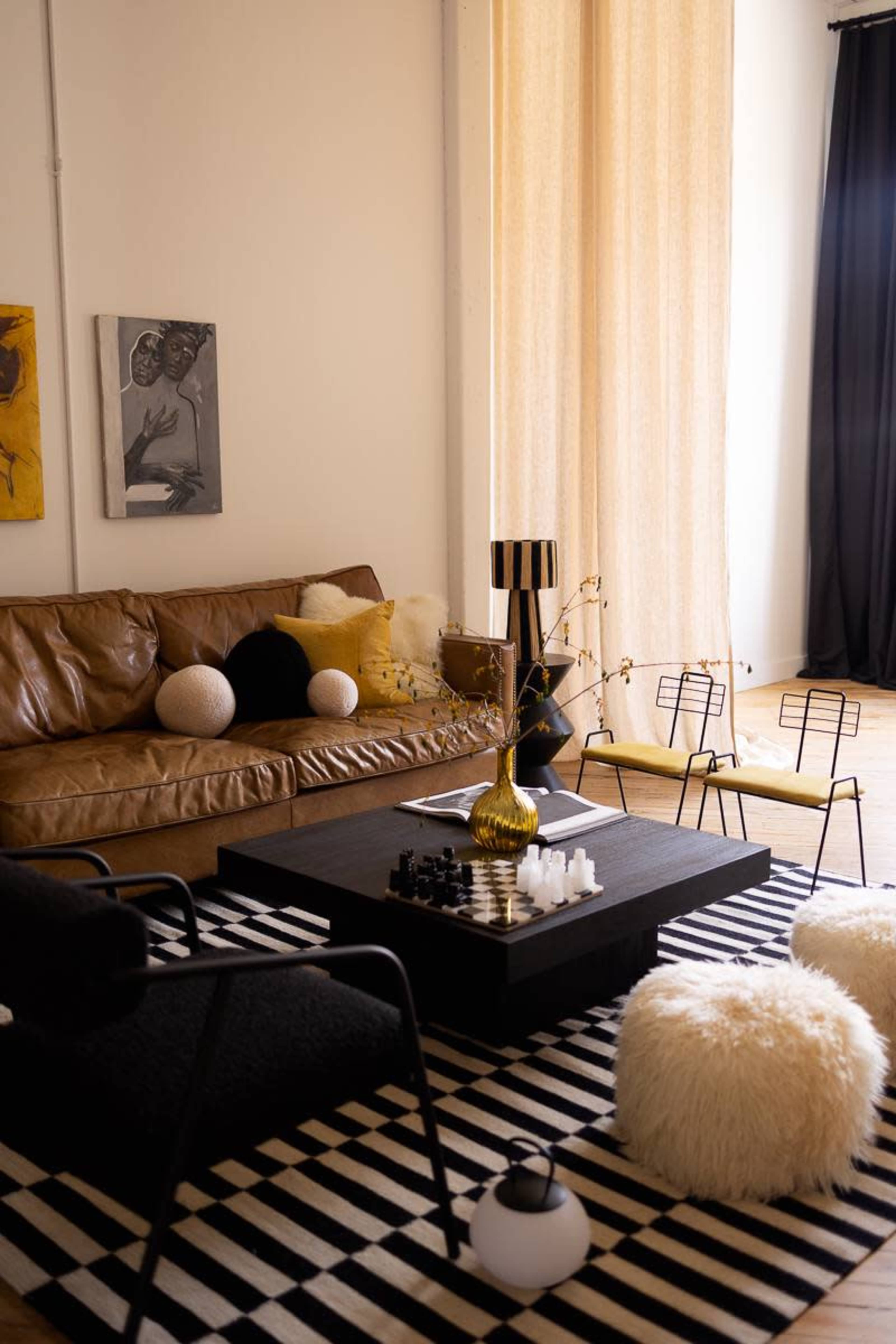 The image shows a stylish living room featuring a brown leather sofa, a black coffee table with a chess set, and various decorative pillows on a striped rug.