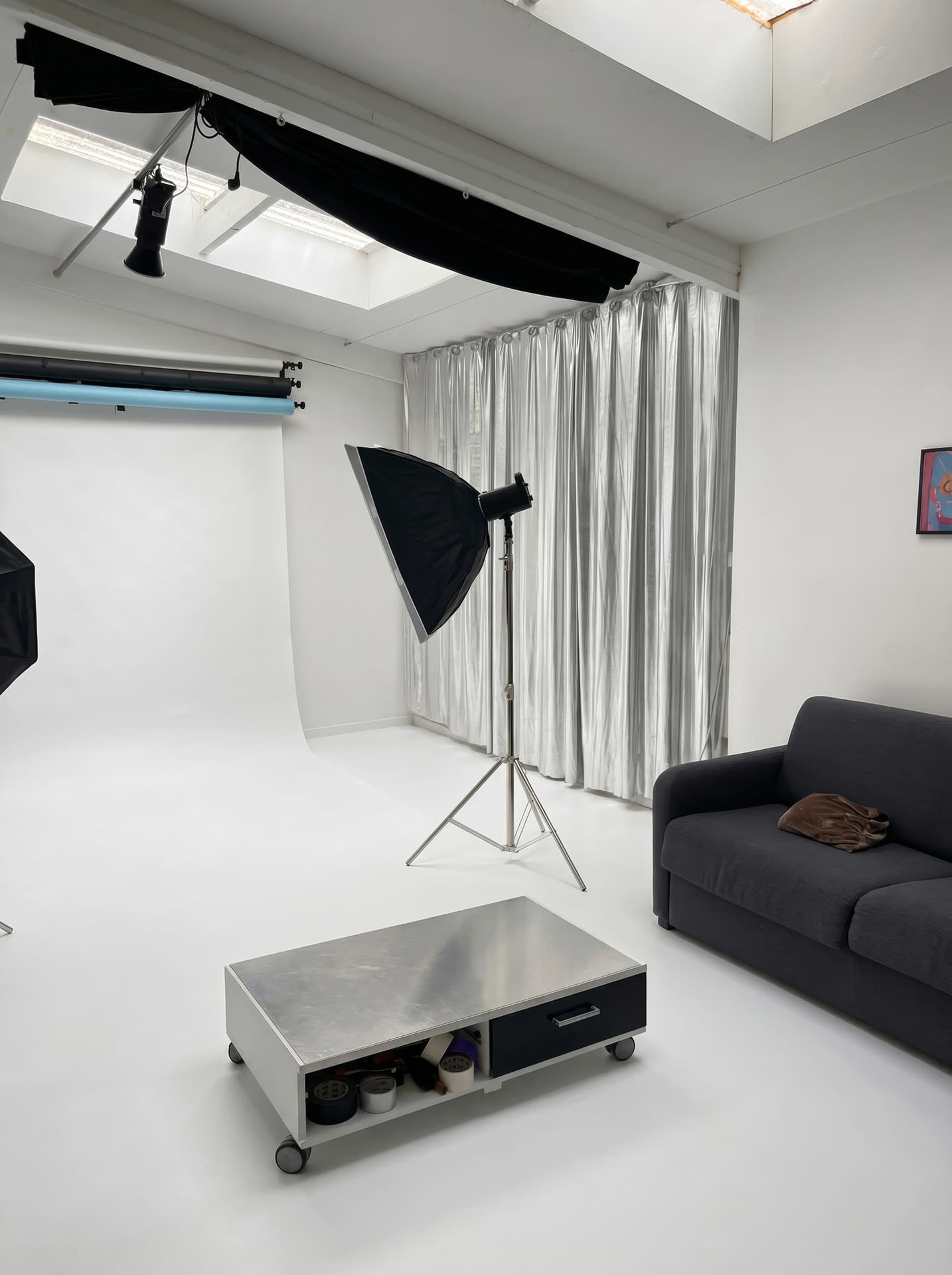 The image shows a photography studio with a white backdrop, a light stand, and a dark gray couch beside a small, wheeled coffee table.