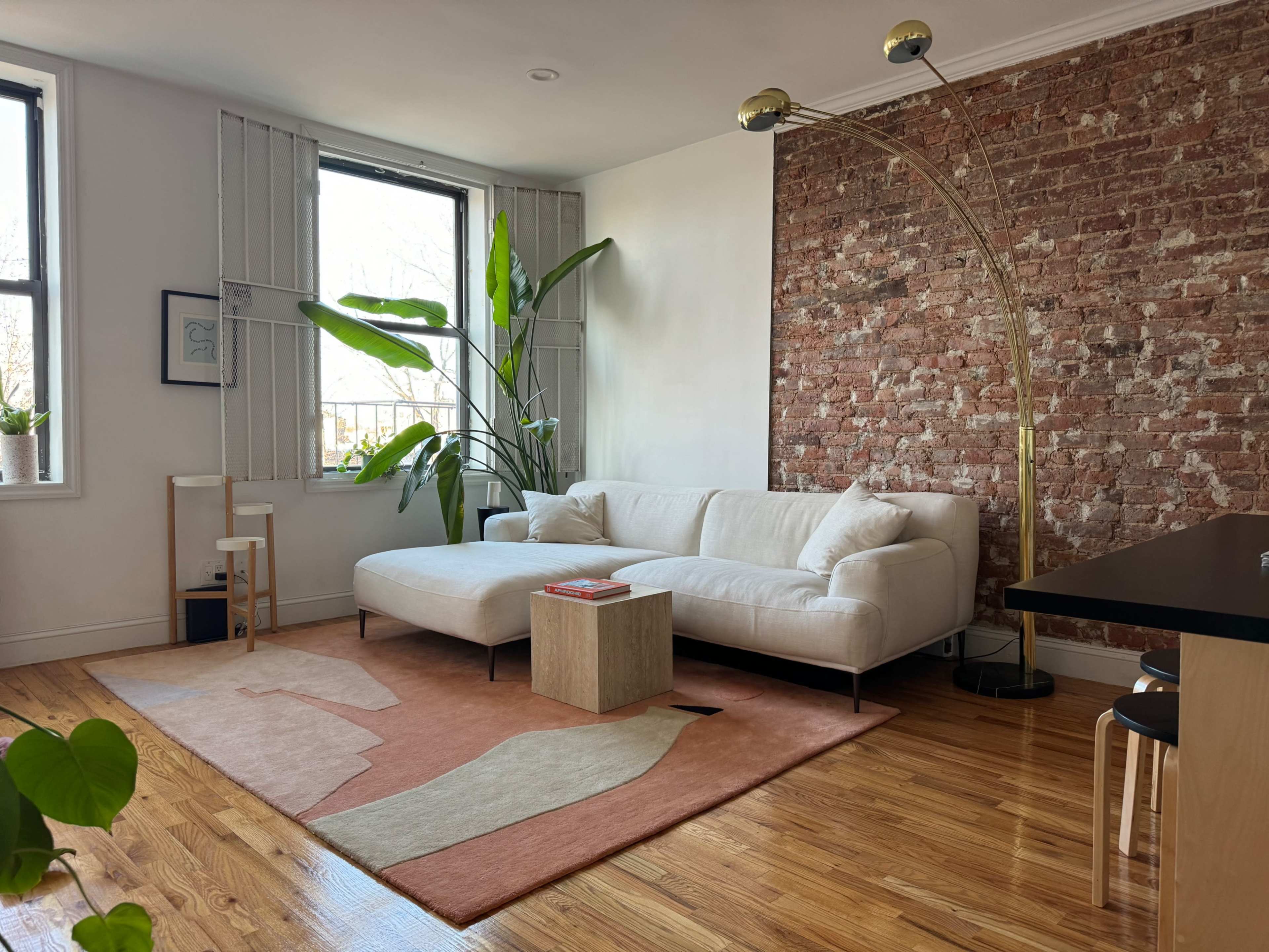 The image shows a modern living room with a white sectional sofa, a wooden coffee table on a patterned rug, and large windows letting in natural light, alongside exposed brick walls and green plants.