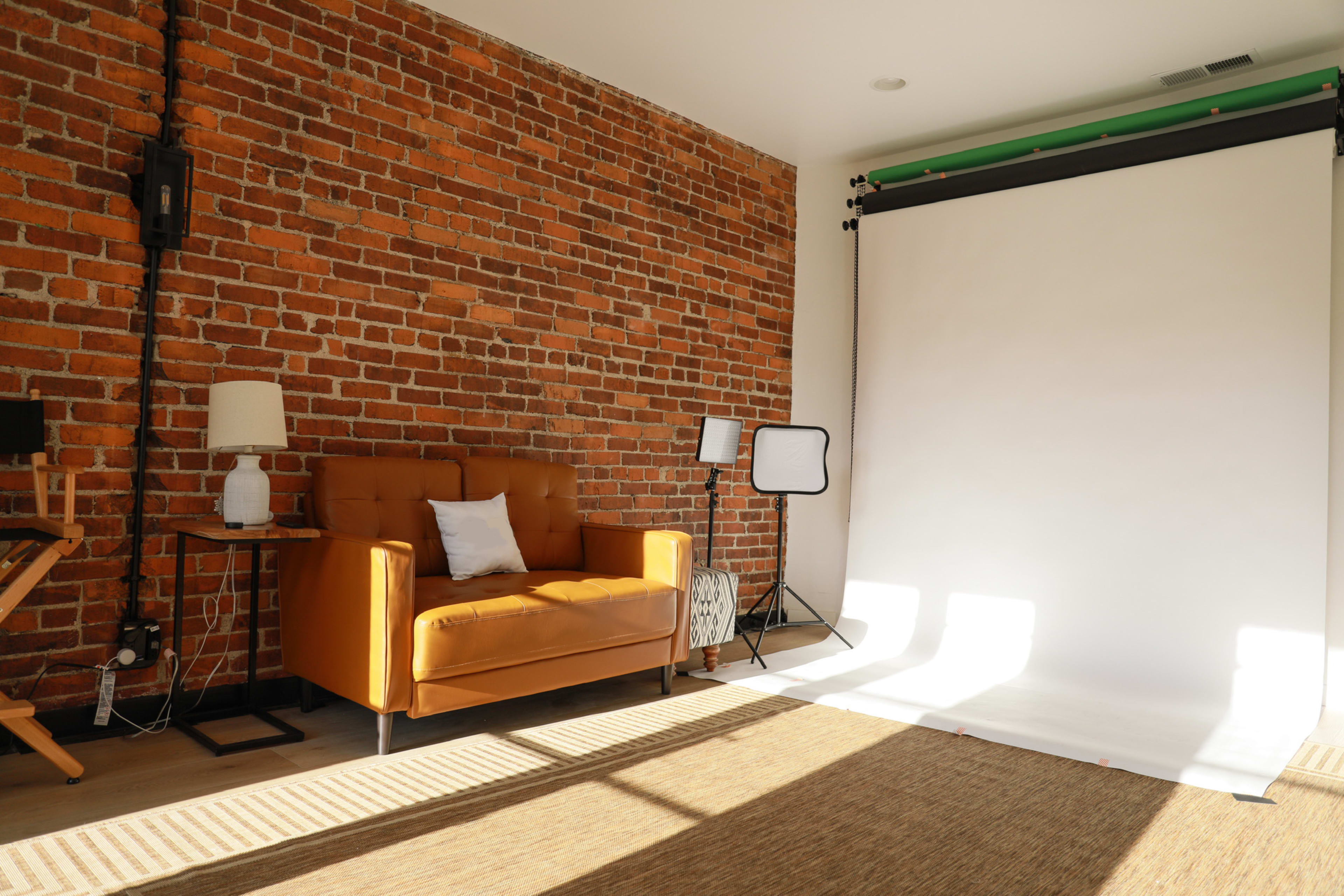 A modern photography studio with a brown leather couch, a white backdrop, and softbox lights against a brick wall.