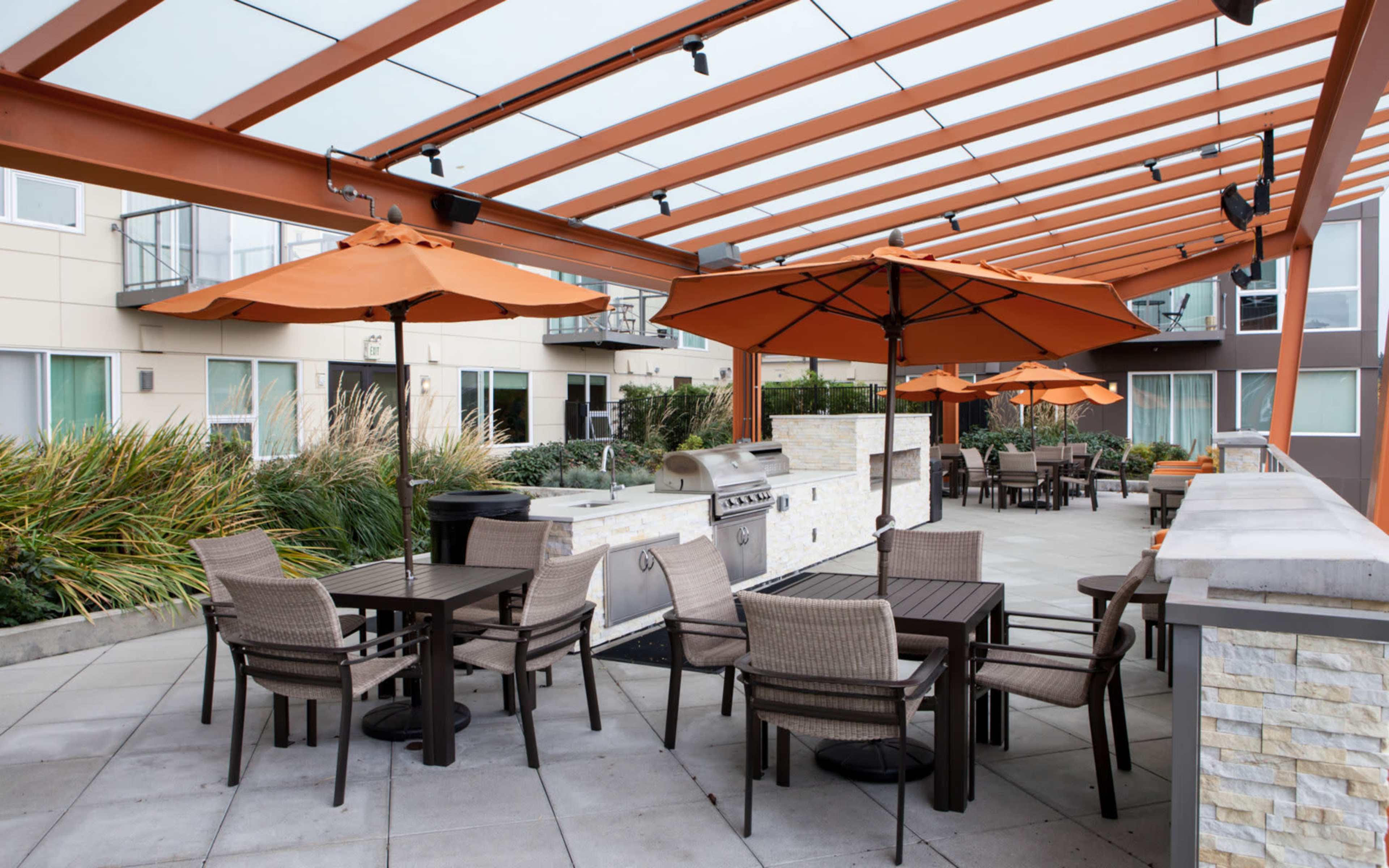 An outdoor dining area with tables and chairs under orange umbrellas, next to a grill and surrounded by modern apartment buildings.